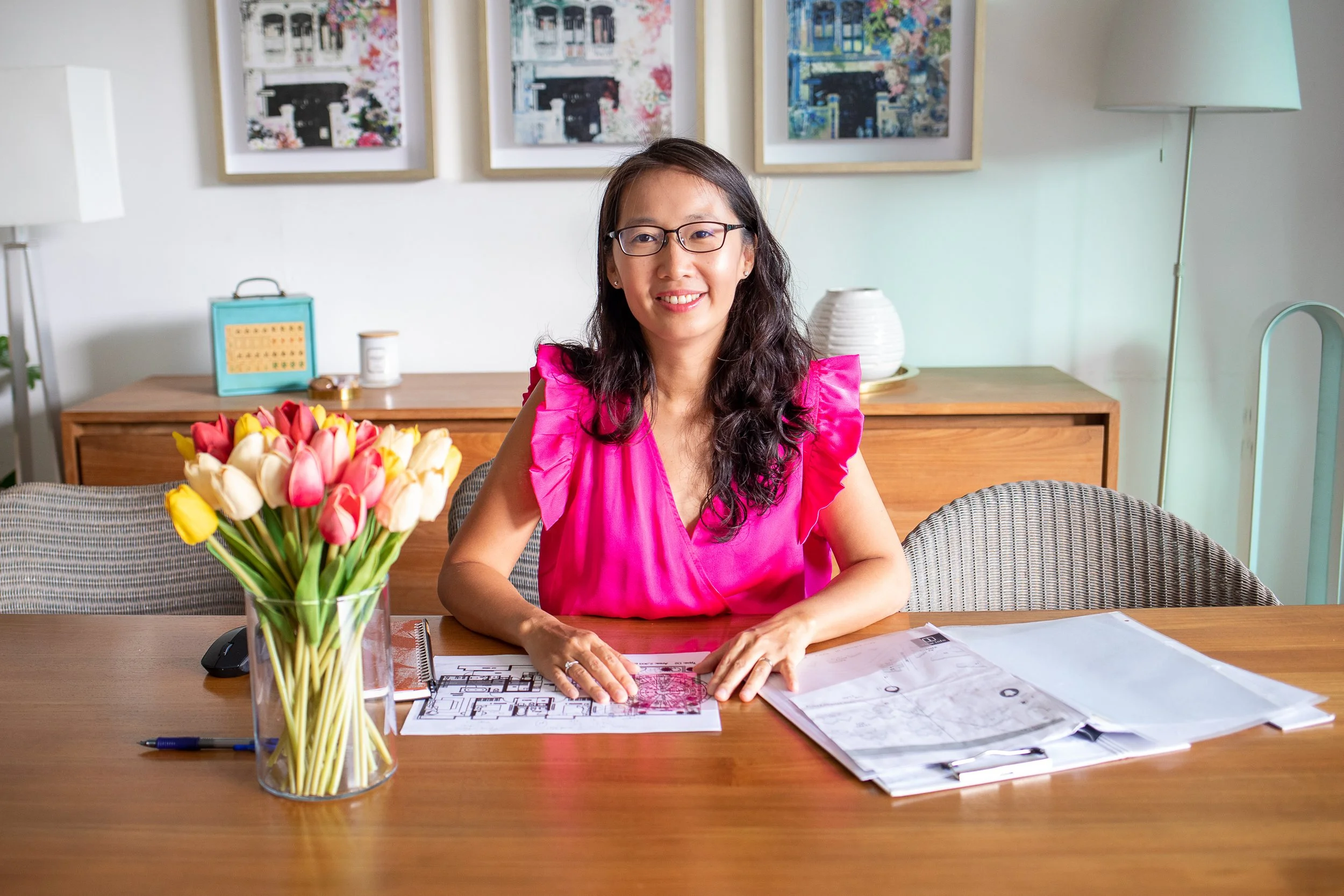 A woman with glasses wearing a pink dress sitting at a wooden table with architectural plans and documents, in a room with framed artwork, a lamp, a vase of tulips, and a sideboard with decorative objects.