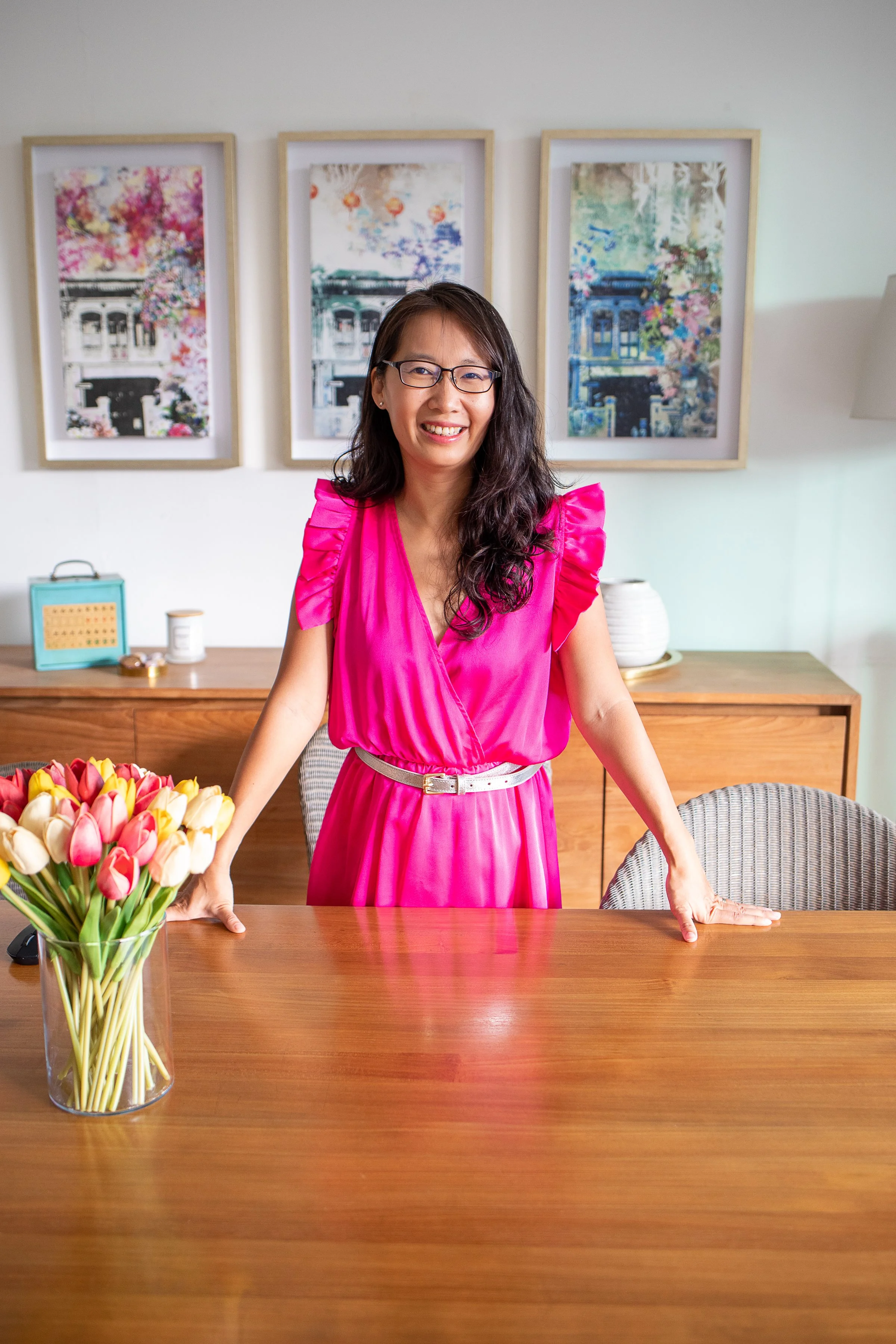 A woman in a bright pink dress standing behind a wooden table with her hands on it. A vase of pink, white, and yellow tulips is on the table. In the background, there are three framed pictures on the wall, a sideboard with decorative items, and a lamp.