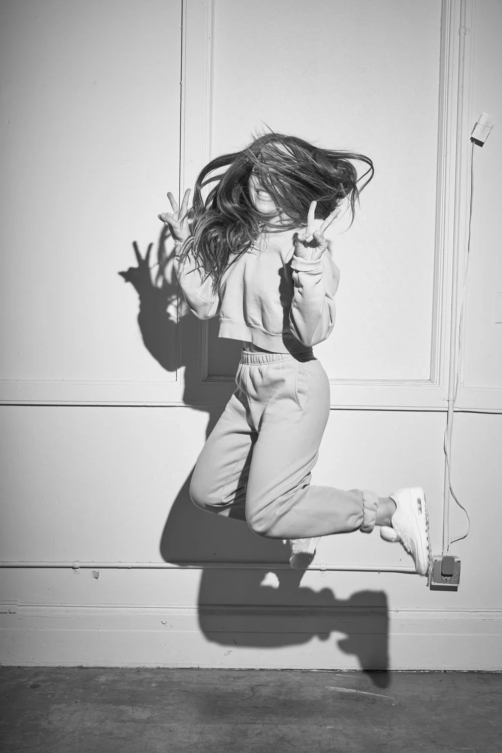 Beth jumping in front of a plain wall, with her hair covering her face, making peace signs with both hands.