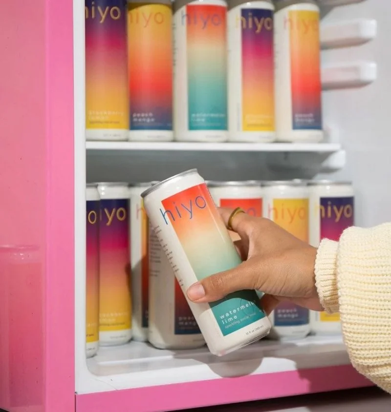 Person holding a Hiya can in front of a fridge filled with similar cans featuring colorful gradient labels.