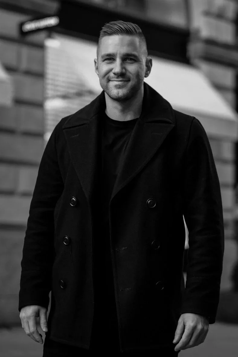 A smiling man with short hair and a beard stands outdoors in front of a brick building, wearing a dark coat.