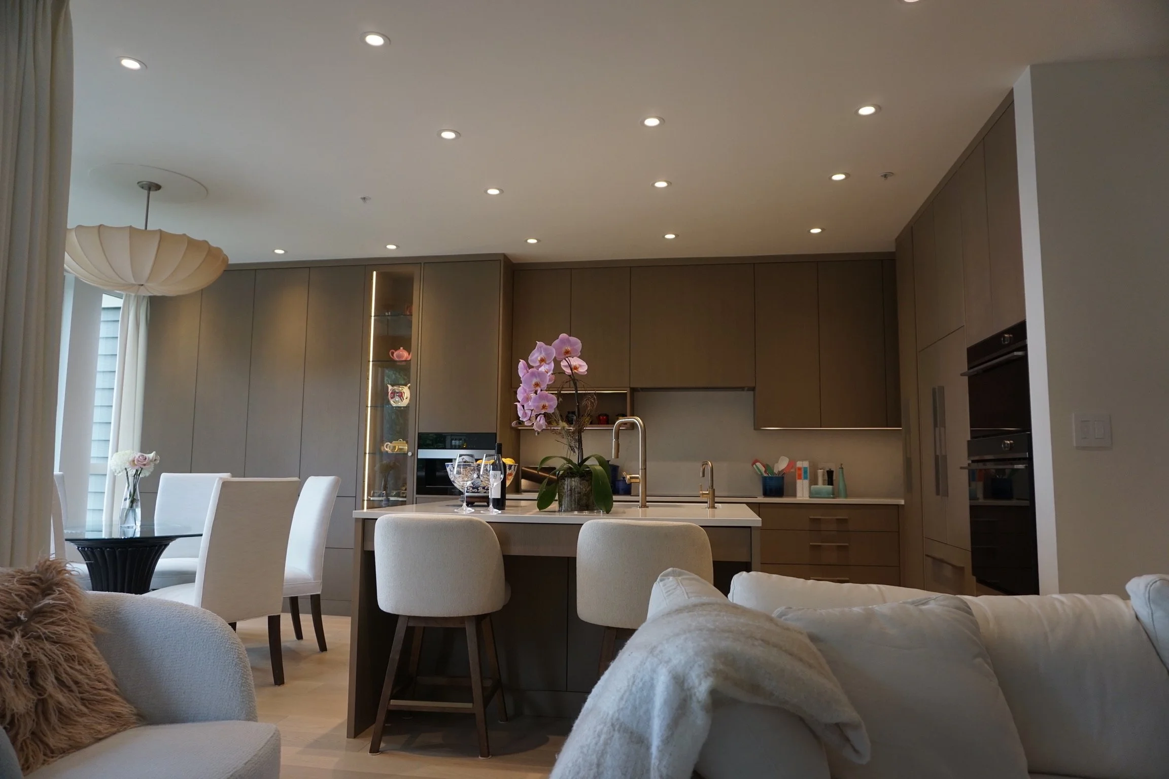 Modern open-concept kitchen with a white island, purple orchids, and bar stools, adjacent to a living space with white chairs and a glass dining table, in a well-lit room with recessed ceiling lights and cabinetry.