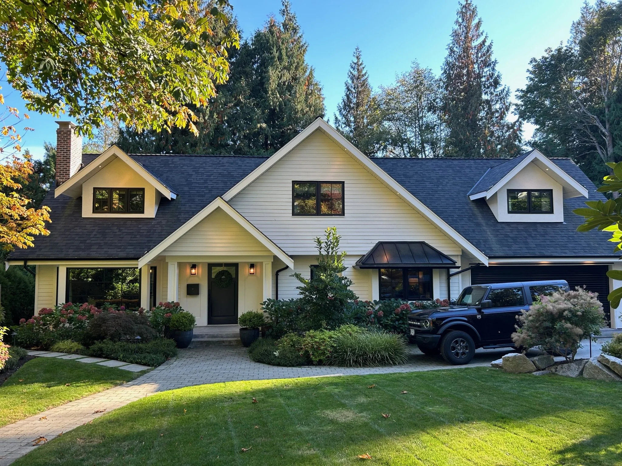 Bright yellow house with black window frames, front porch, landscaped yard, surrounding trees, cloudy sky, dark-colored car in driveway.
