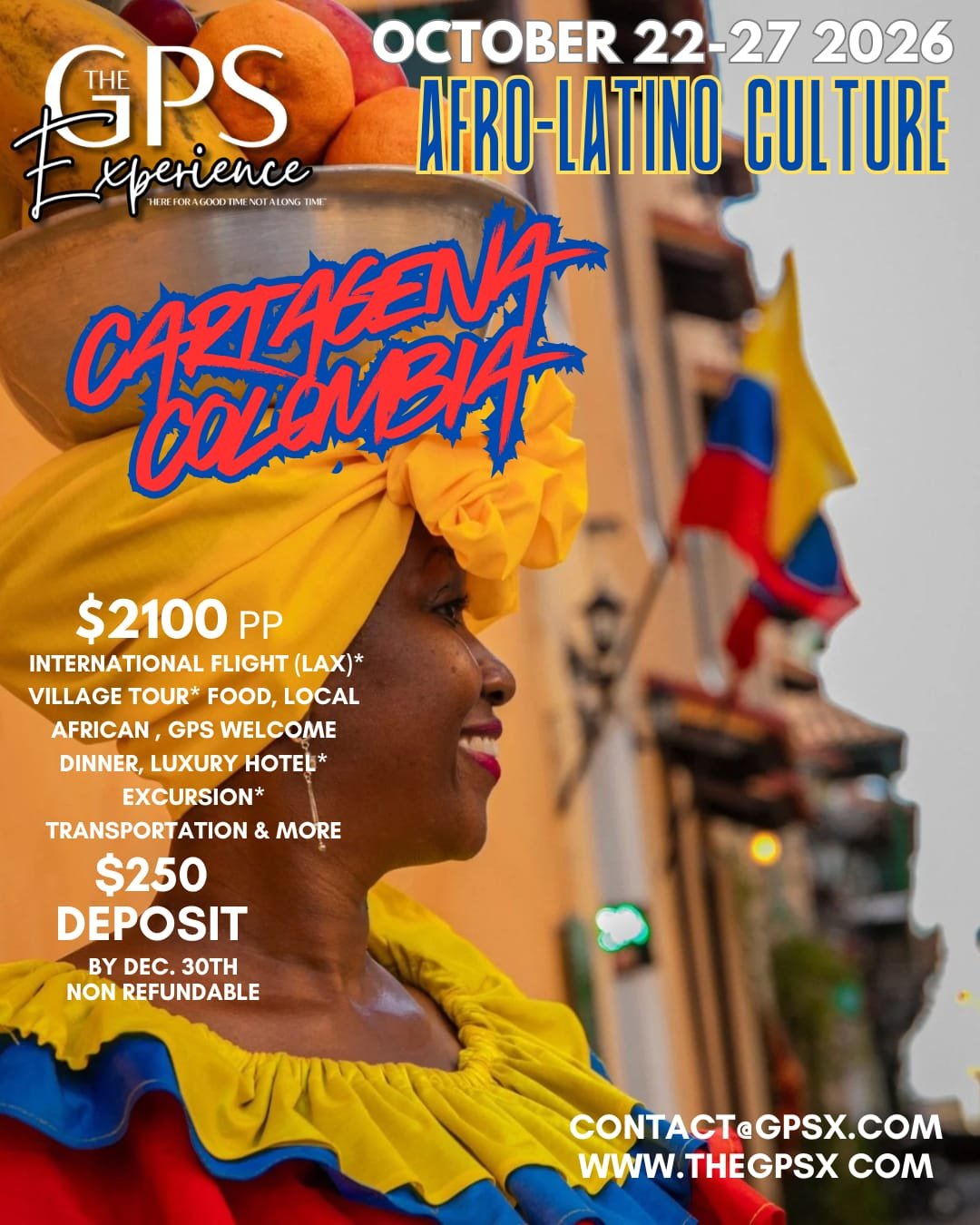 A woman wearing a vibrant yellow head wrap and colorful clothing, smiling, with a bowl of fruit on her head in a street scene with a blurry flag in the background.