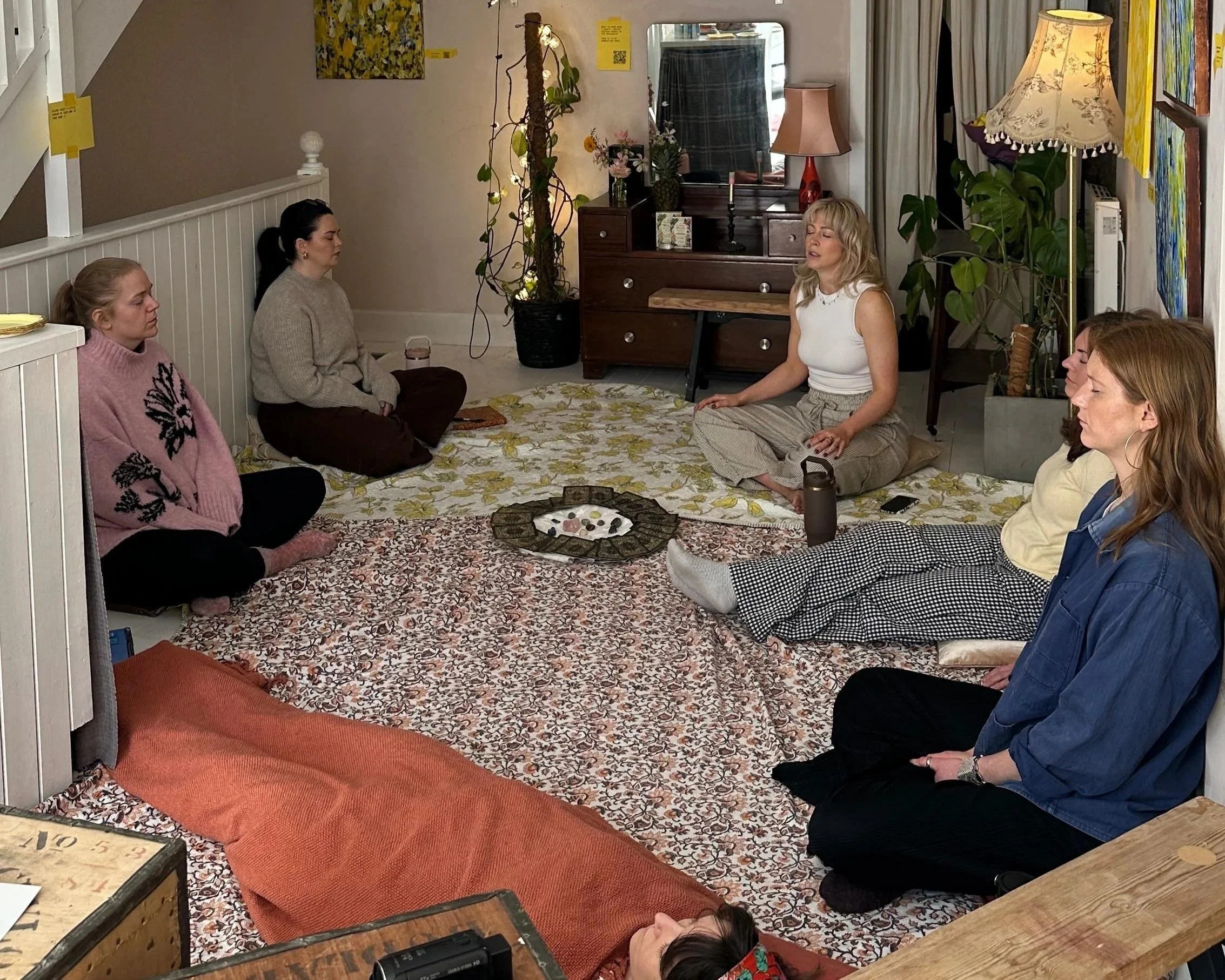 A group of women participating in a meditation or mindfulness session in a cozy living room, sitting on rugs with a small table holding stones in the center, surrounded by plants, artwork, and a cabinet with a mirror.