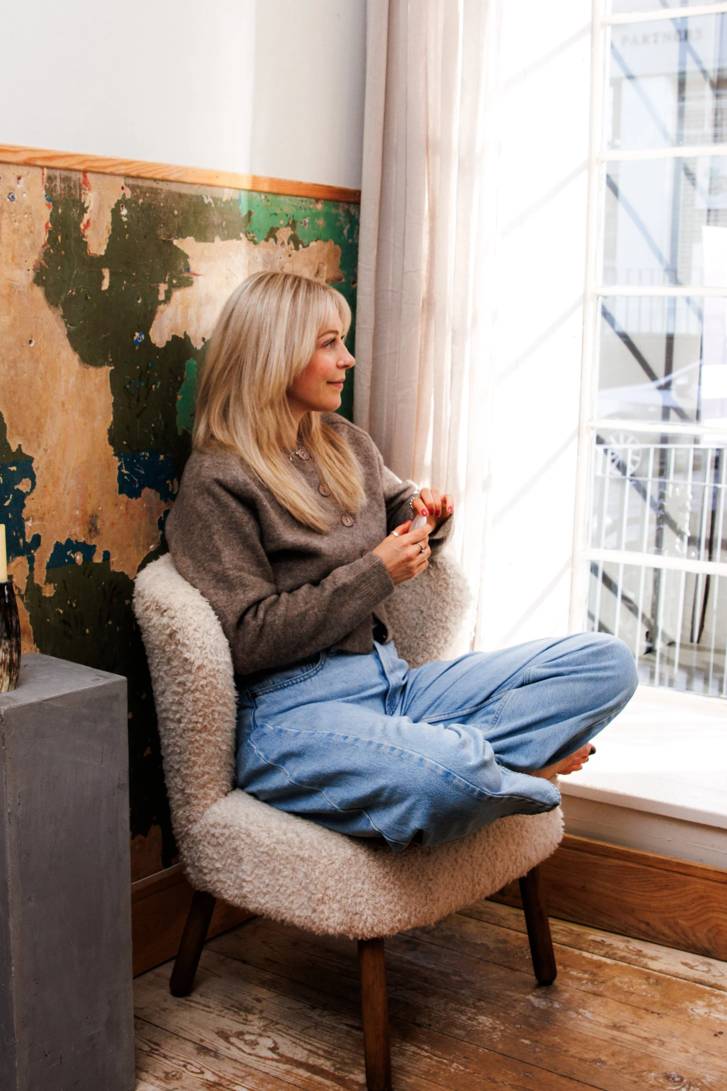 A woman with blonde hair sitting cross-legged on a cozy, cream-colored chair by a large window with natural light. She appears relaxed, holding a smartphone, wearing a brown sweater and blue jeans. The interior features a wooden floor and a partially visible old world map on the wall.