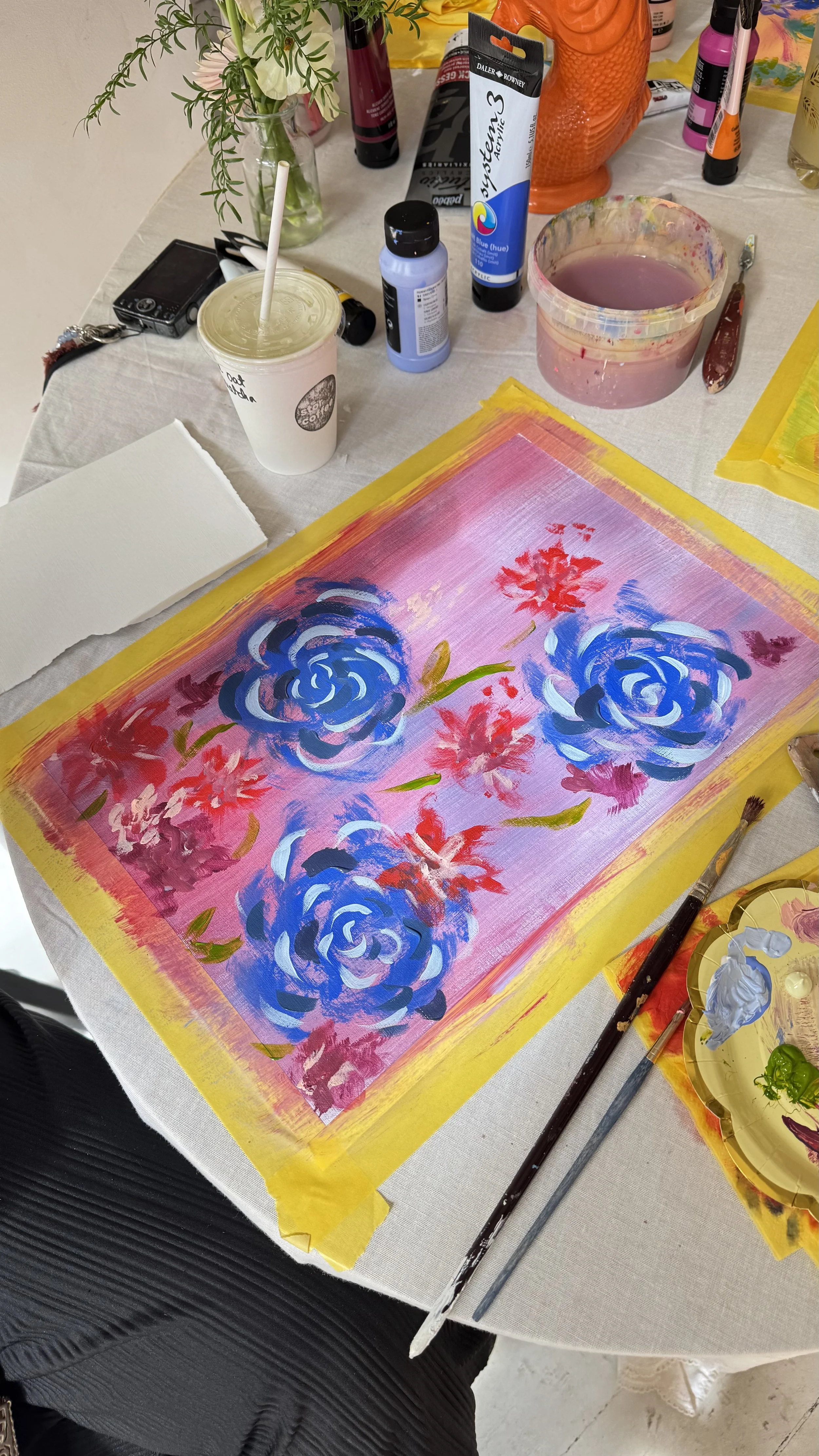 A colorful floral painting in progress on a table covered with painting supplies, including brushes, paints, and a palette. The table has a white cloth and the artwork features large blue and pink roses with red flowers on a pink background outlined with yellow tape.