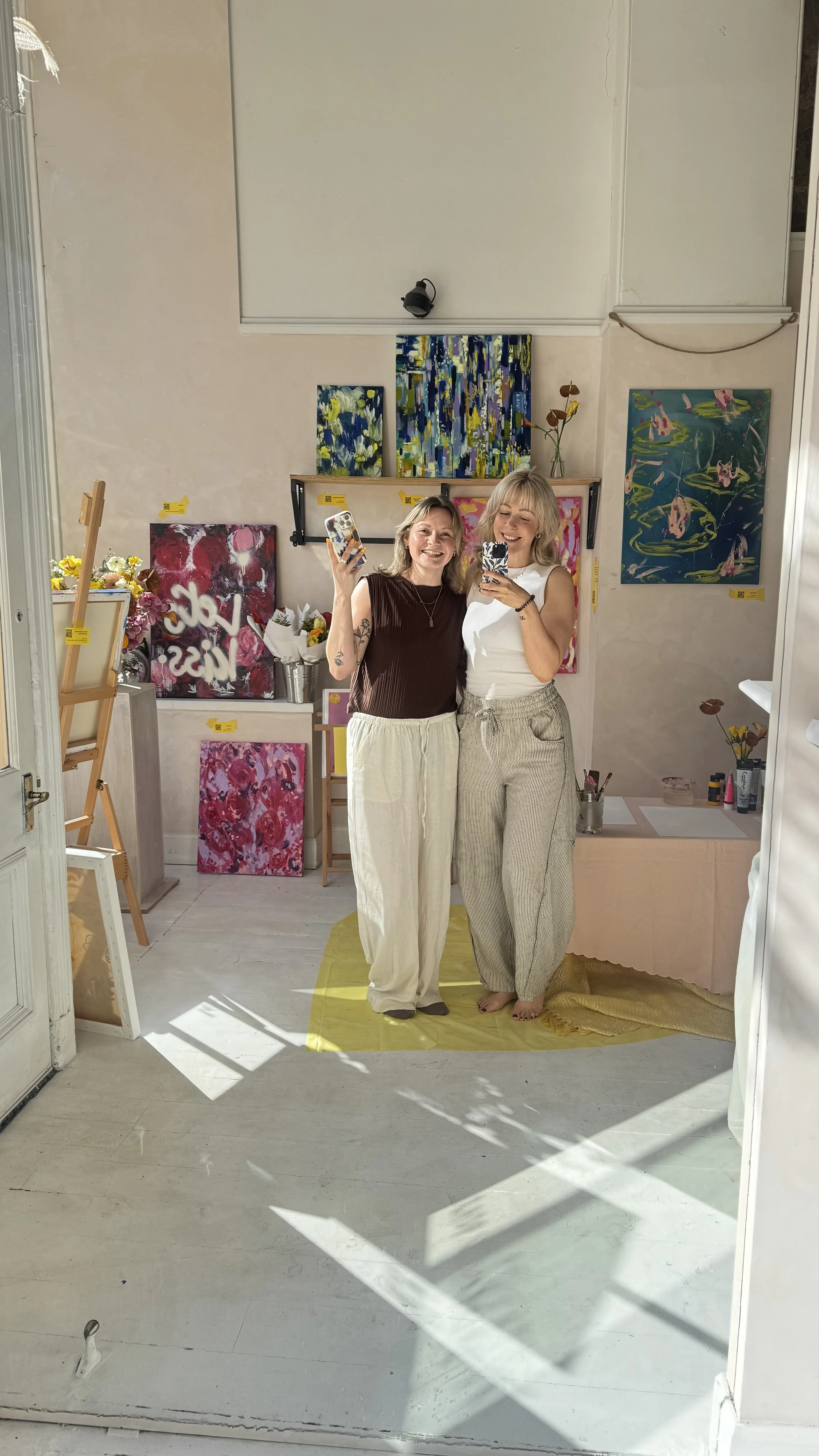 Two women standing in an art studio, smiling and taking a selfie. The studio has colorful paintings on the wall, with flowers and art supplies around.