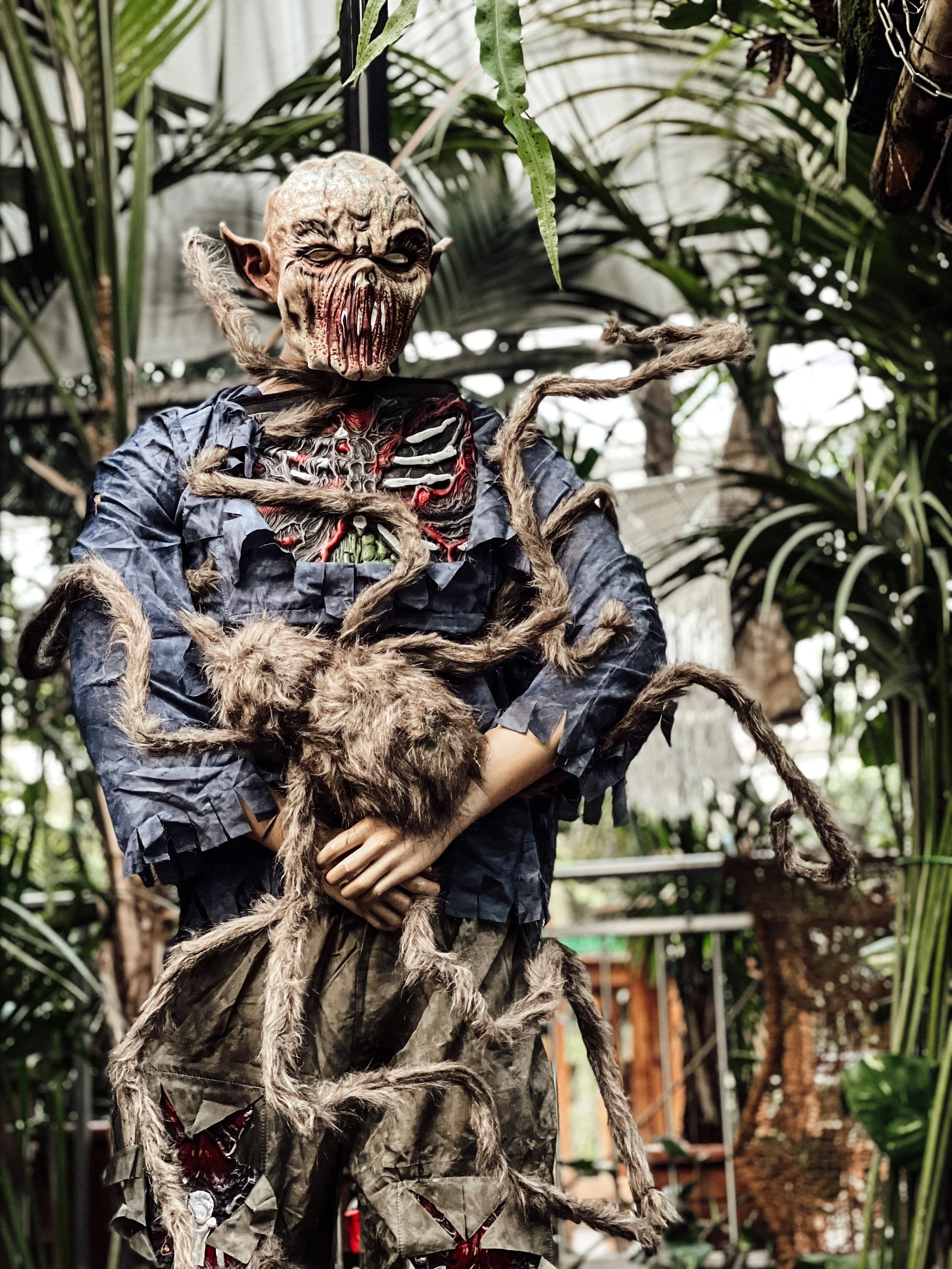 Gruselige Halloween-Dekoration mit Monsterfigur in einem Garten, umgeben von Pflanzen. Die Figur hat einen deformierten Kopf und Tentakel, die aus der Brust wachsen.