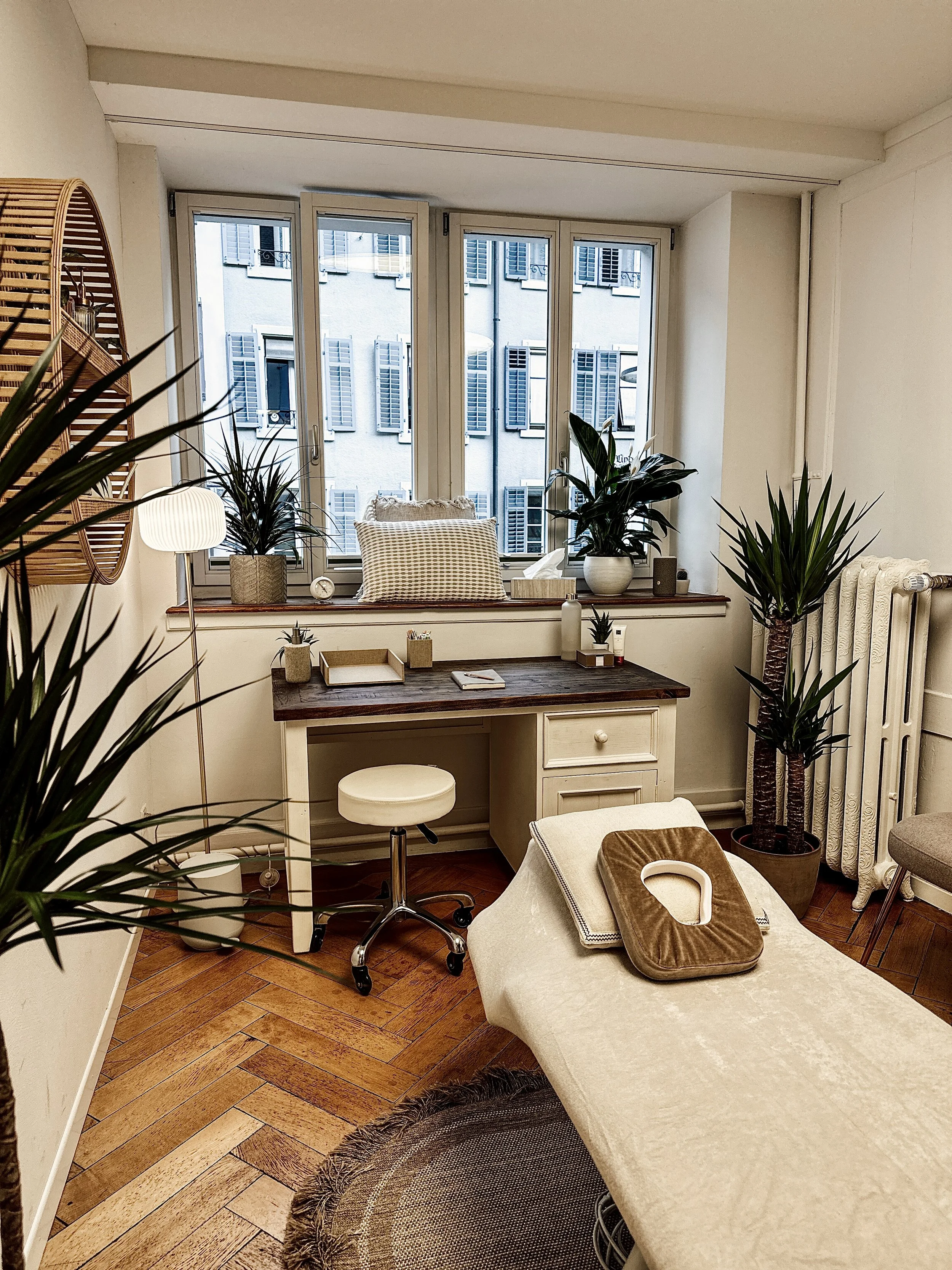 Behandlungsraum mit Massageliege, Holzboden, Schreibtisch, Hocker und Pflanzen; Fenster mit Blick auf Nachbargebäude.