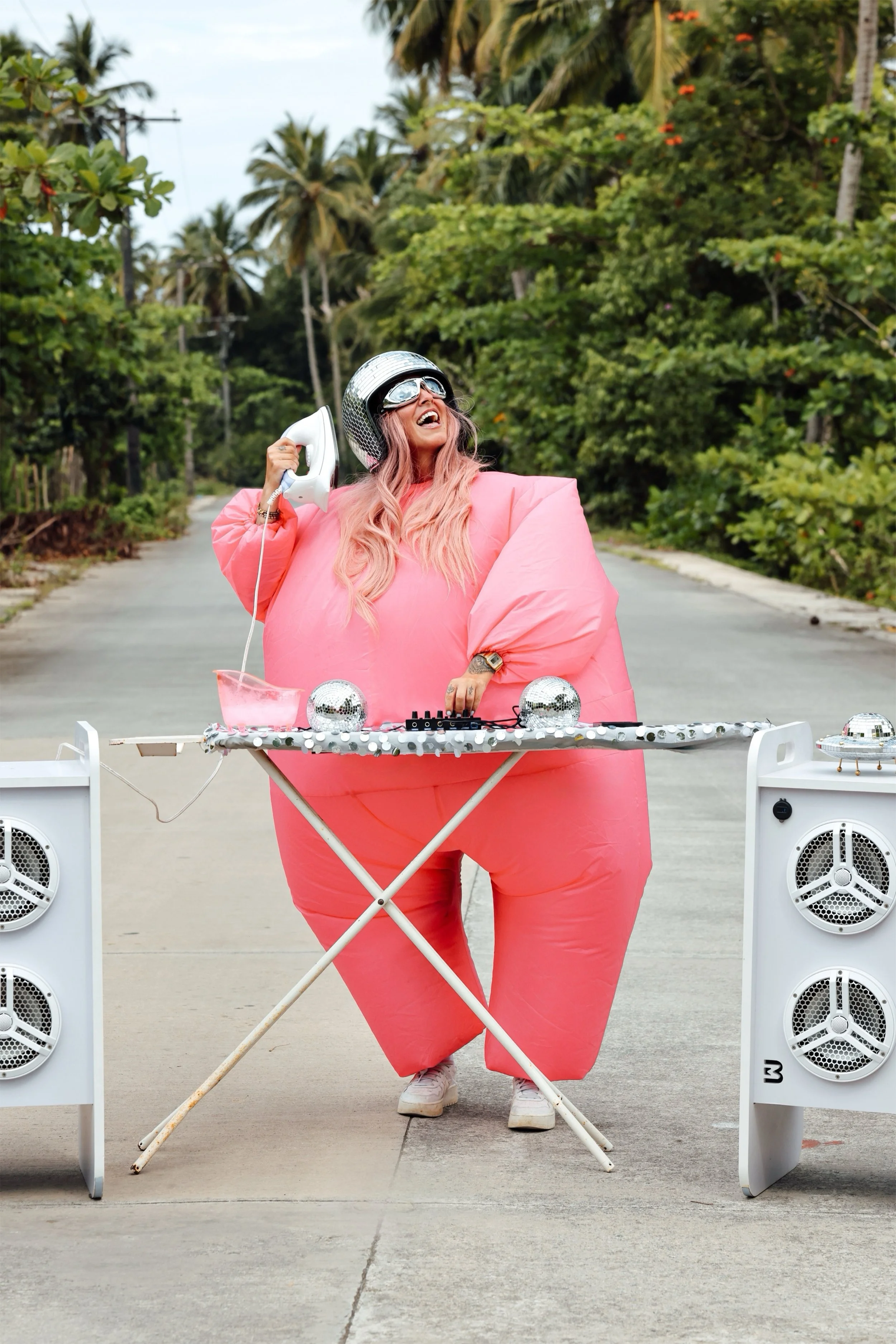 Person in einem aufblasbaren rosa Kostüm mit Helm, spielt Musik auf einem DJ-Pult in einer tropischen Umgebung.