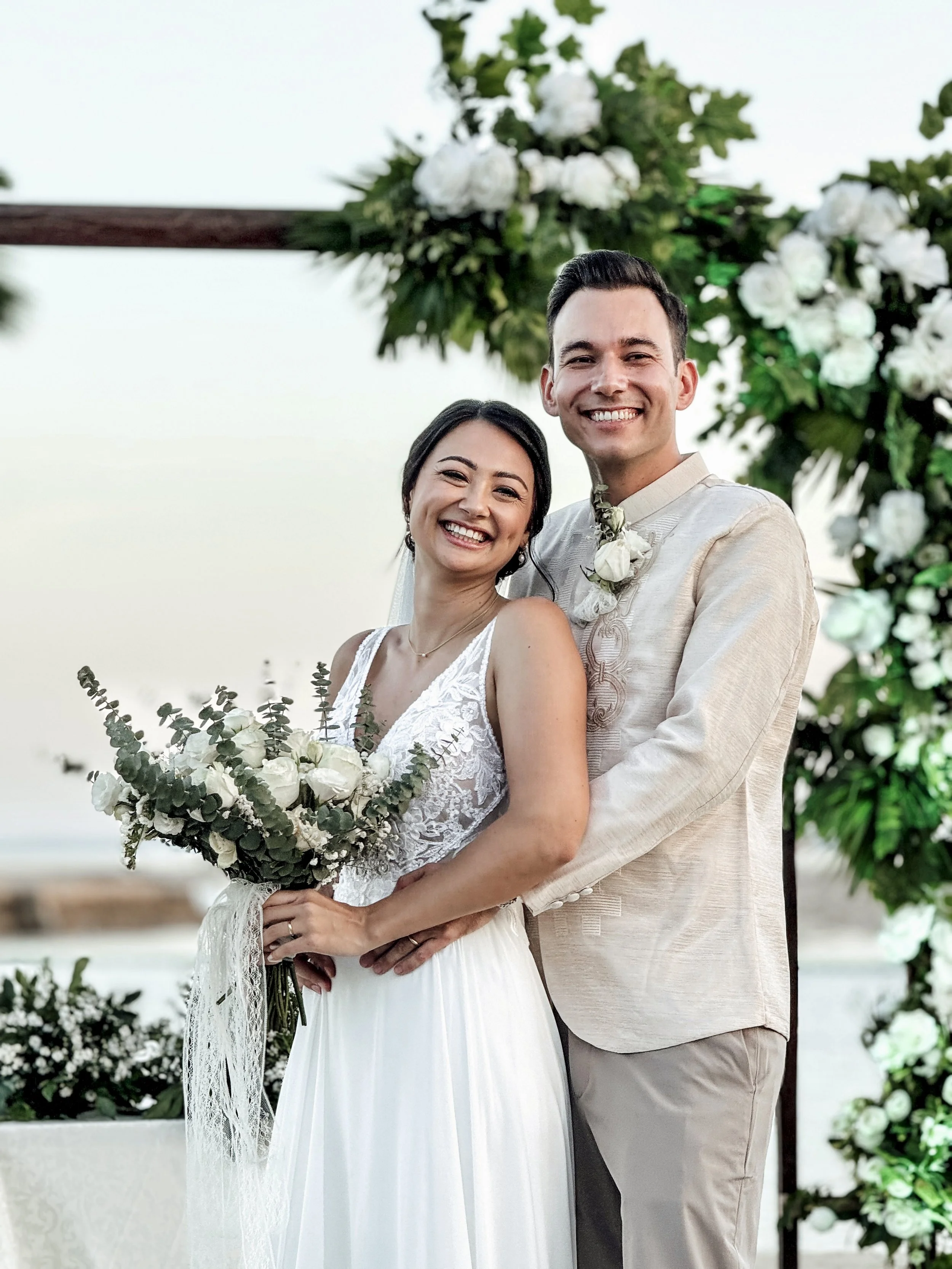 Ein Hochzeitspaar lächelt und posiert in eleganter Kleidung vor einem mit Blumen geschmückten Bogen.