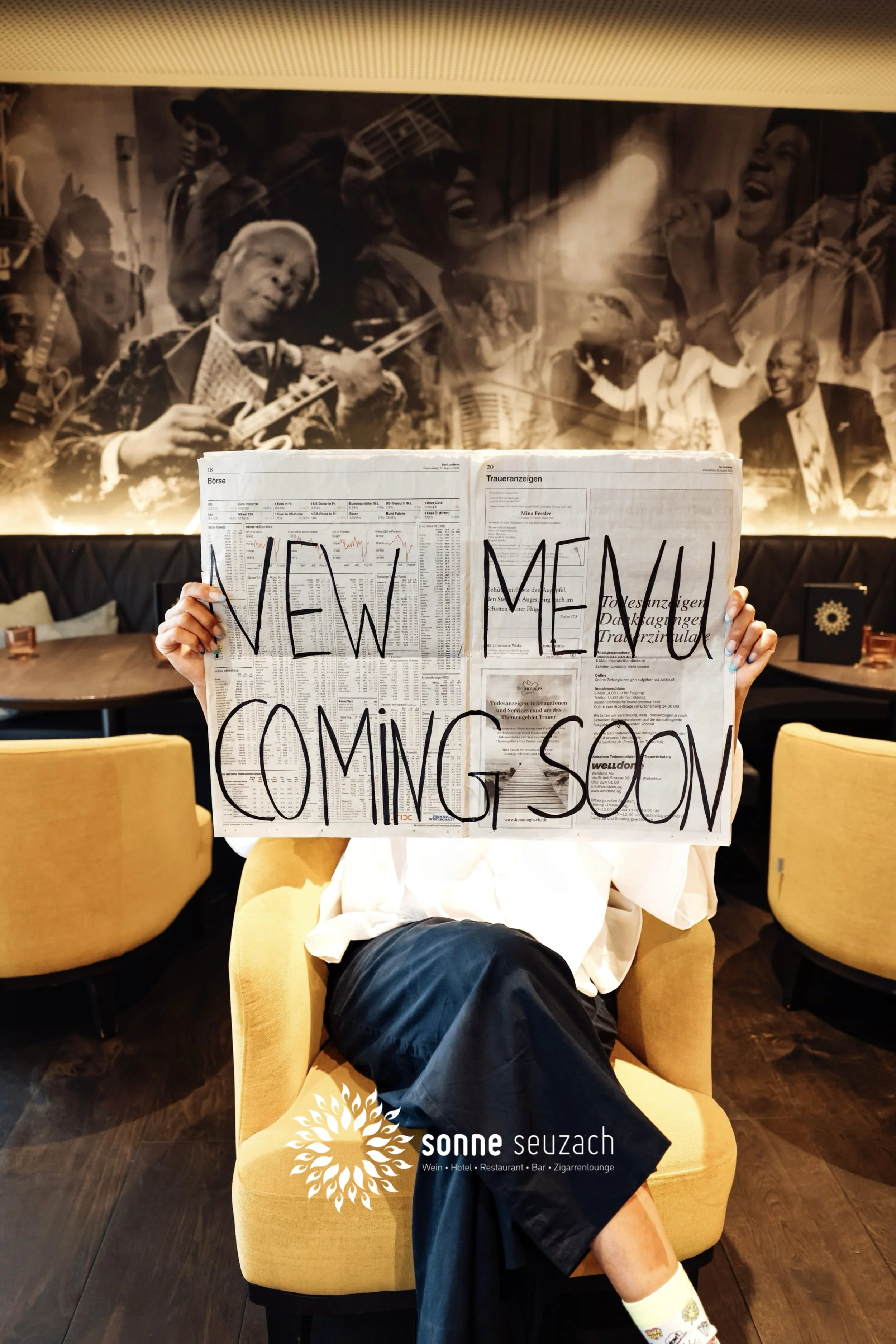 Eine Person sitzt in einem gelben Sessel und hält eine Zeitung vor dem Gesicht mit der großen Aufschrift "NEW MENU COMING SOON". Im Hintergrund ist eine Wand mit schwarzweißen Bildern von Musikern. Unten rechts ist das Logo von "Sonne Seuzach" zu sehen.