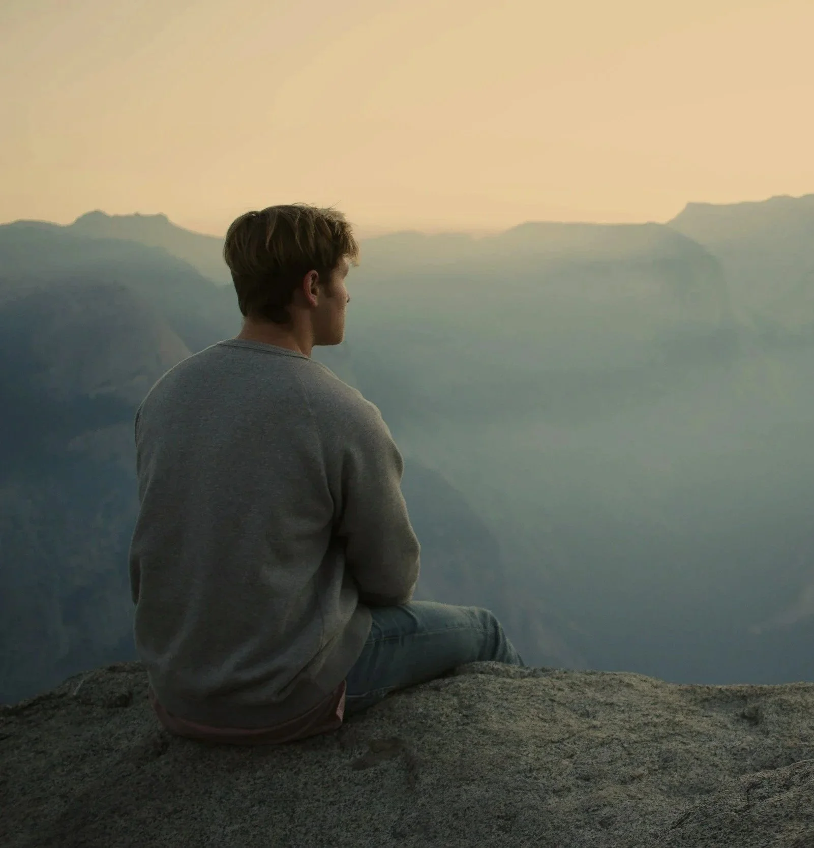 A person sitting on a rocky surface looking out over a mountain landscape at sunset or sunrise.