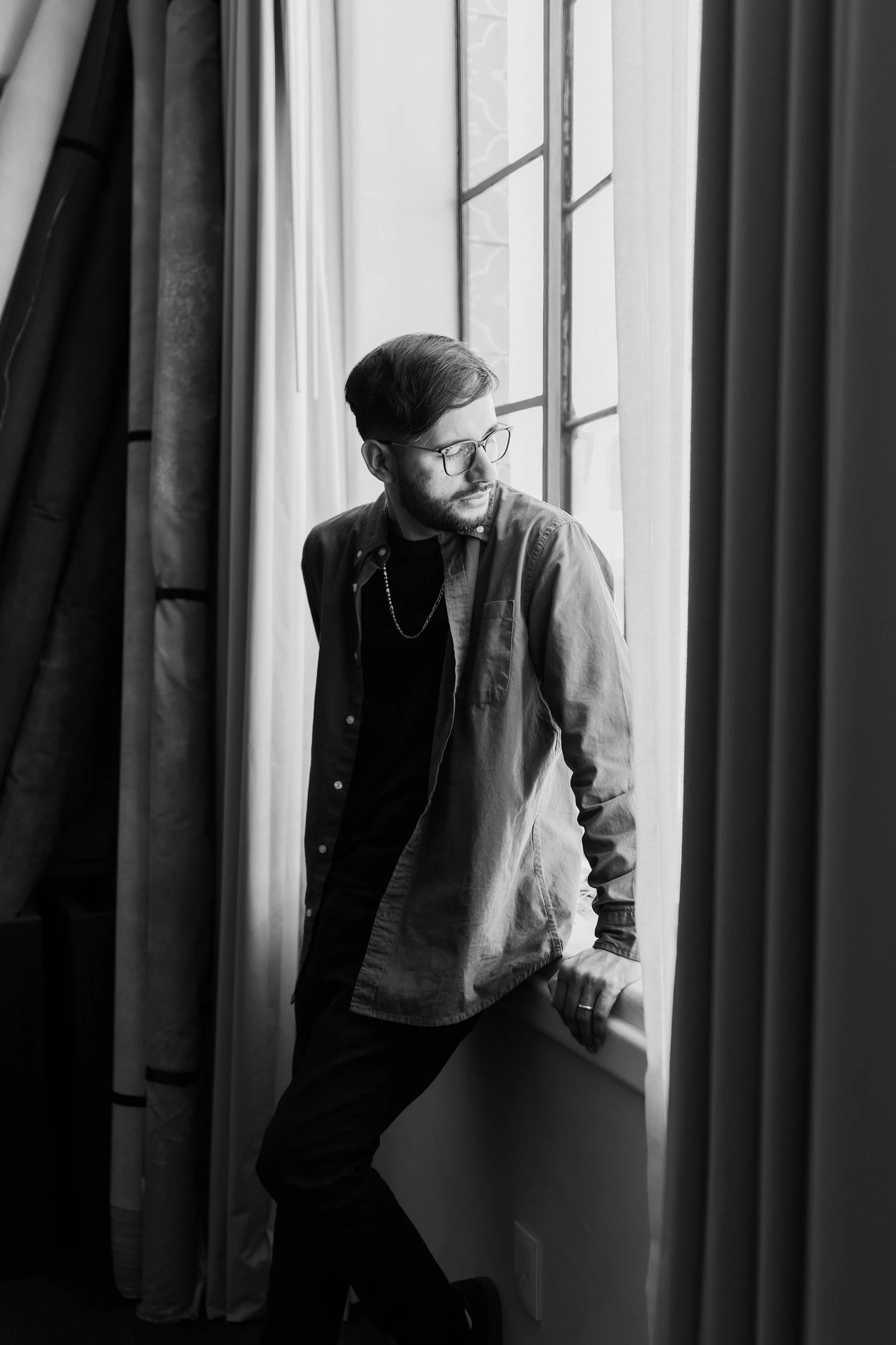A black and white photo of a young man with glasses, wearing a casual shirt and a chain necklace, standing near a window with curtains, looking out.