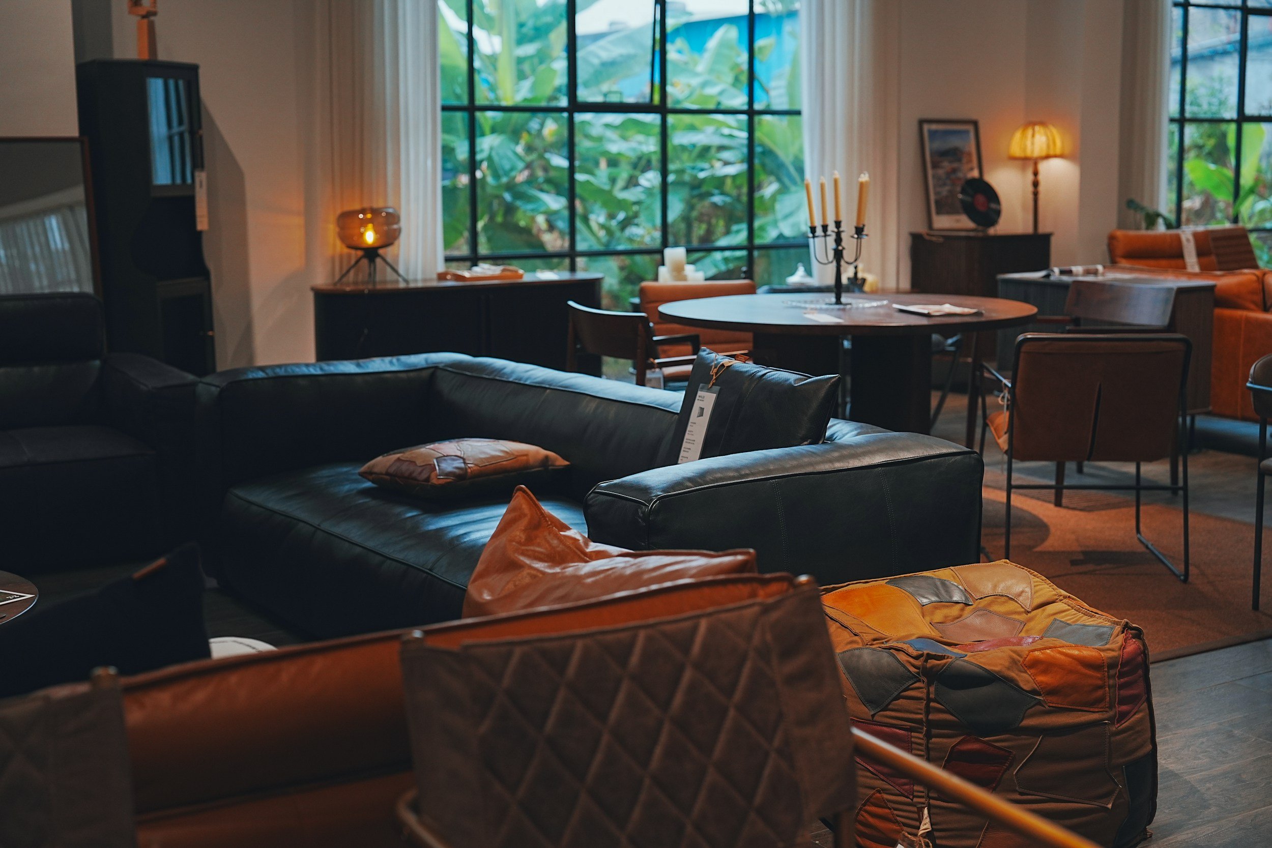 Cozy living room with large windows showing greenery outside, furnished with leather sofas, wooden chairs, and vintage decor like candles, a record player, and framed artwork.