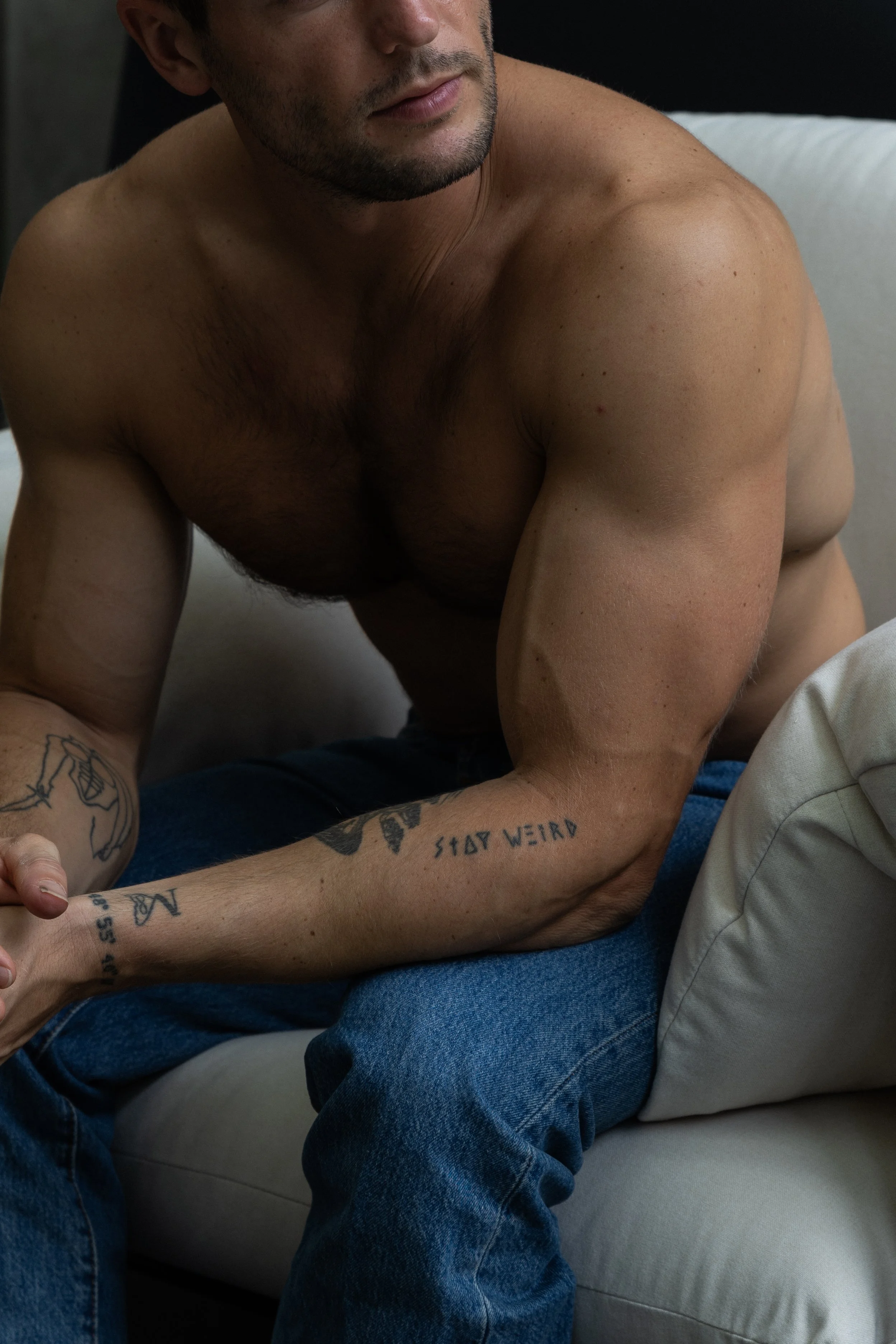 A shirtless man with tattoos on his arms sits on a couch, partially visible, with a woman in light-colored pants next to him. Their faces are cut off from the frame.