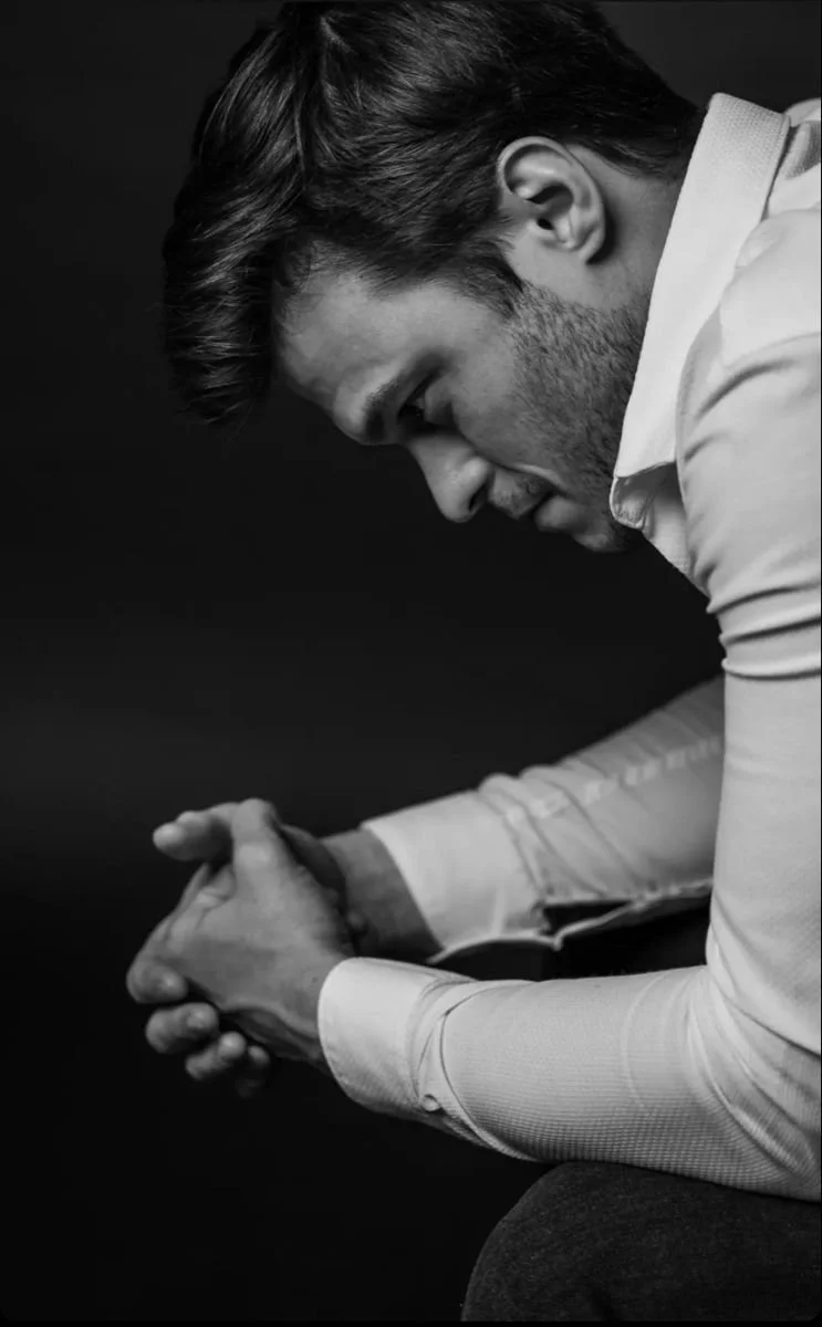Black and white photo of a man with hair styled back, dressed in a white shirt, sitting with his elbows resting on his knees, looking down with hands clasped