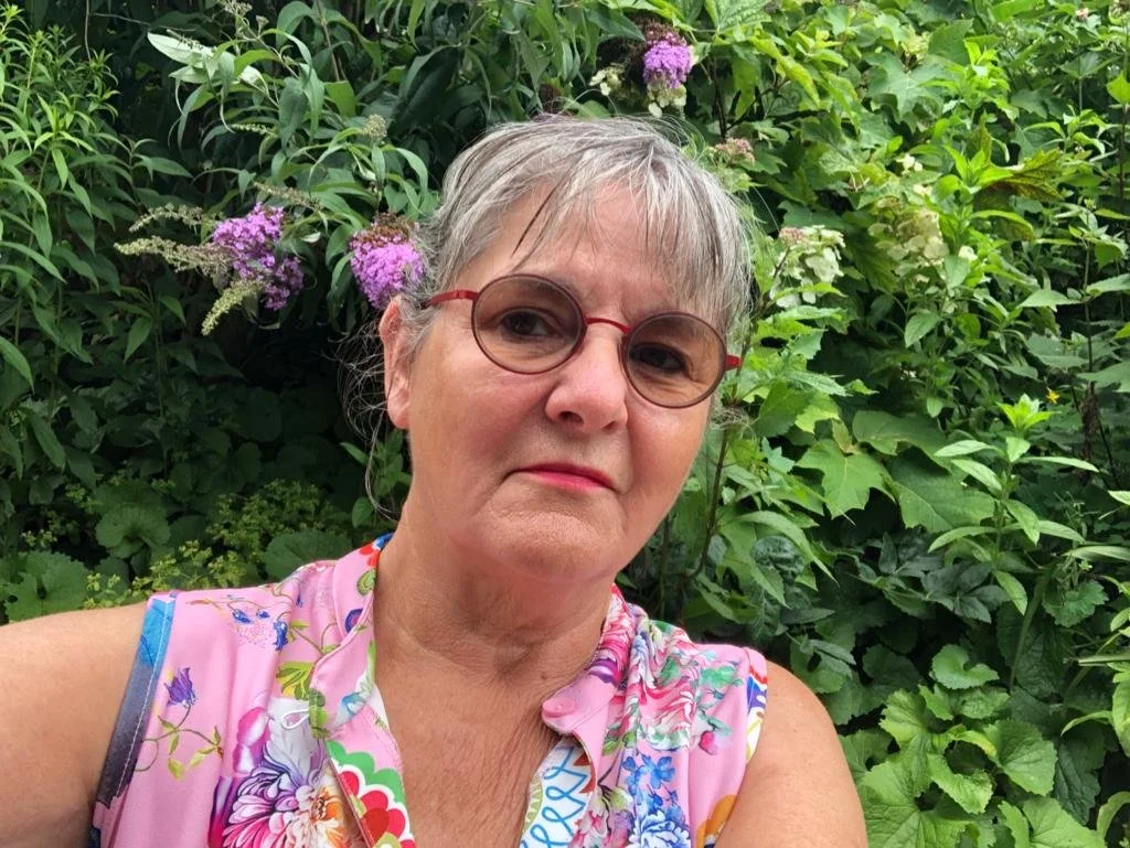 Een oudere vrouw met grijs haar en een roze bloemenprint top, draagt ronde glazen met een rood montuur, staat voor een groene tuin met bloemen.