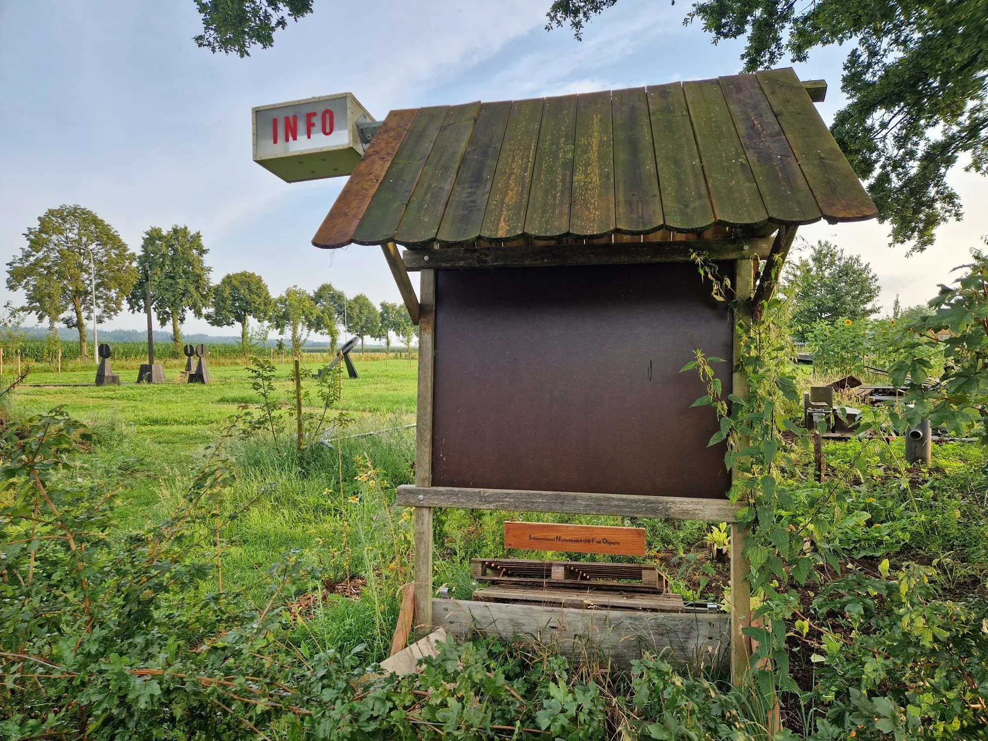 Een leeg informatiebord met een houten structuur in een groene tuin, met een klein rood bordje onderaan en een rood en wit bord met de tekst "INFO" bovenaan links.