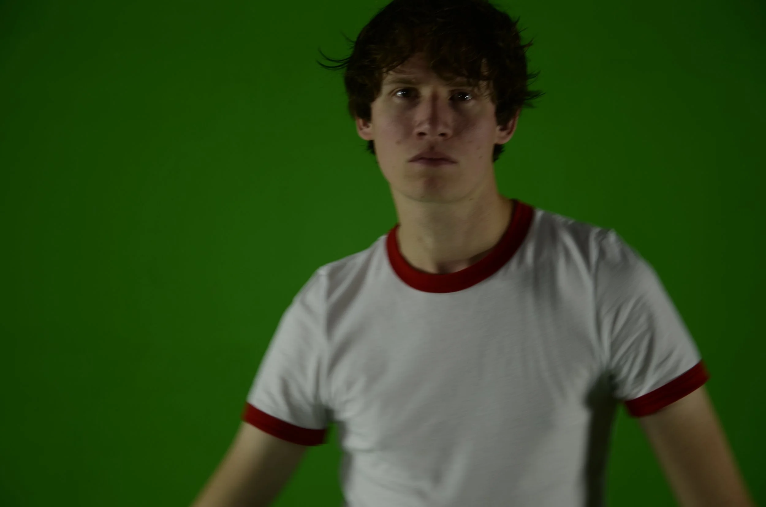 Portret van een jonge man met warrig bruin haar, gekleed in een wit T-shirt met rode boord, tegen een groene achtergrond.