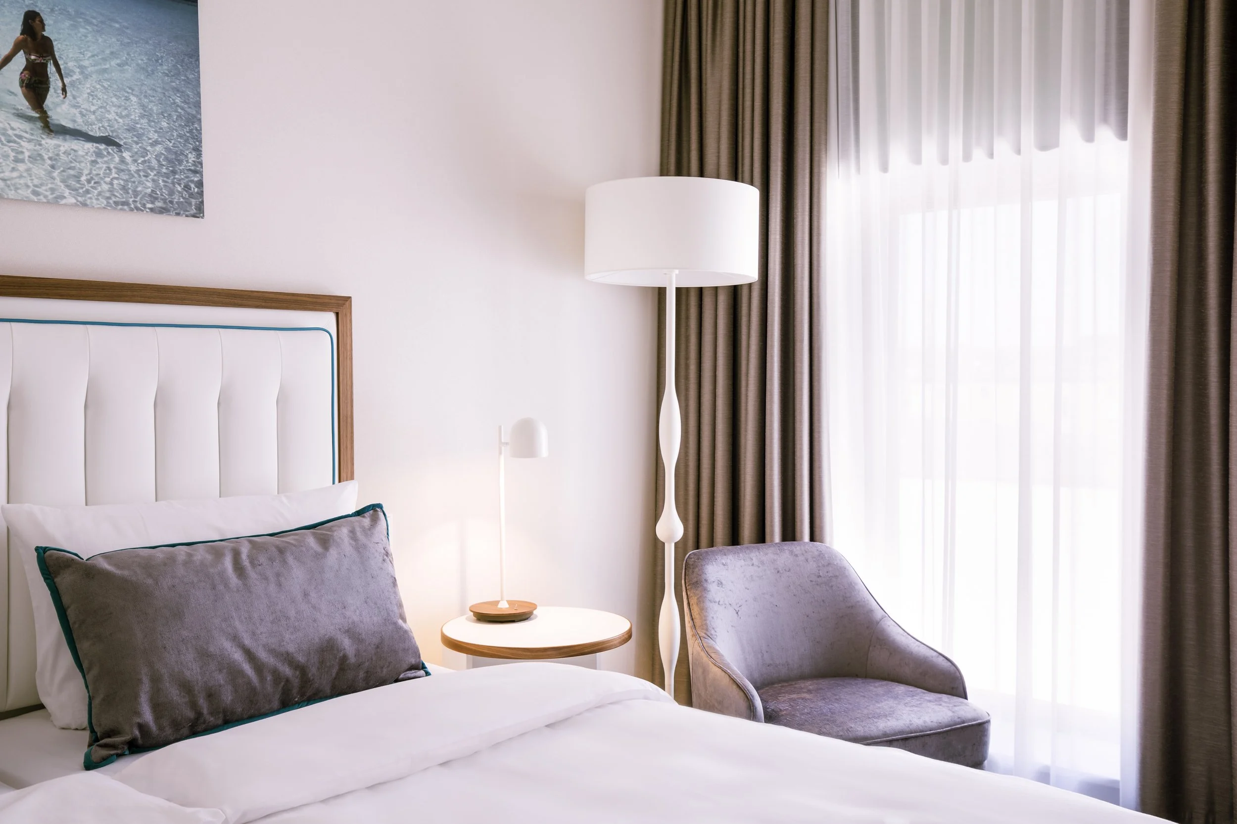 A bedroom with a bed, gray pillows, and white bedding, beside a side table with a white lamp, a tall white floor lamp, and a window with beige curtains.
