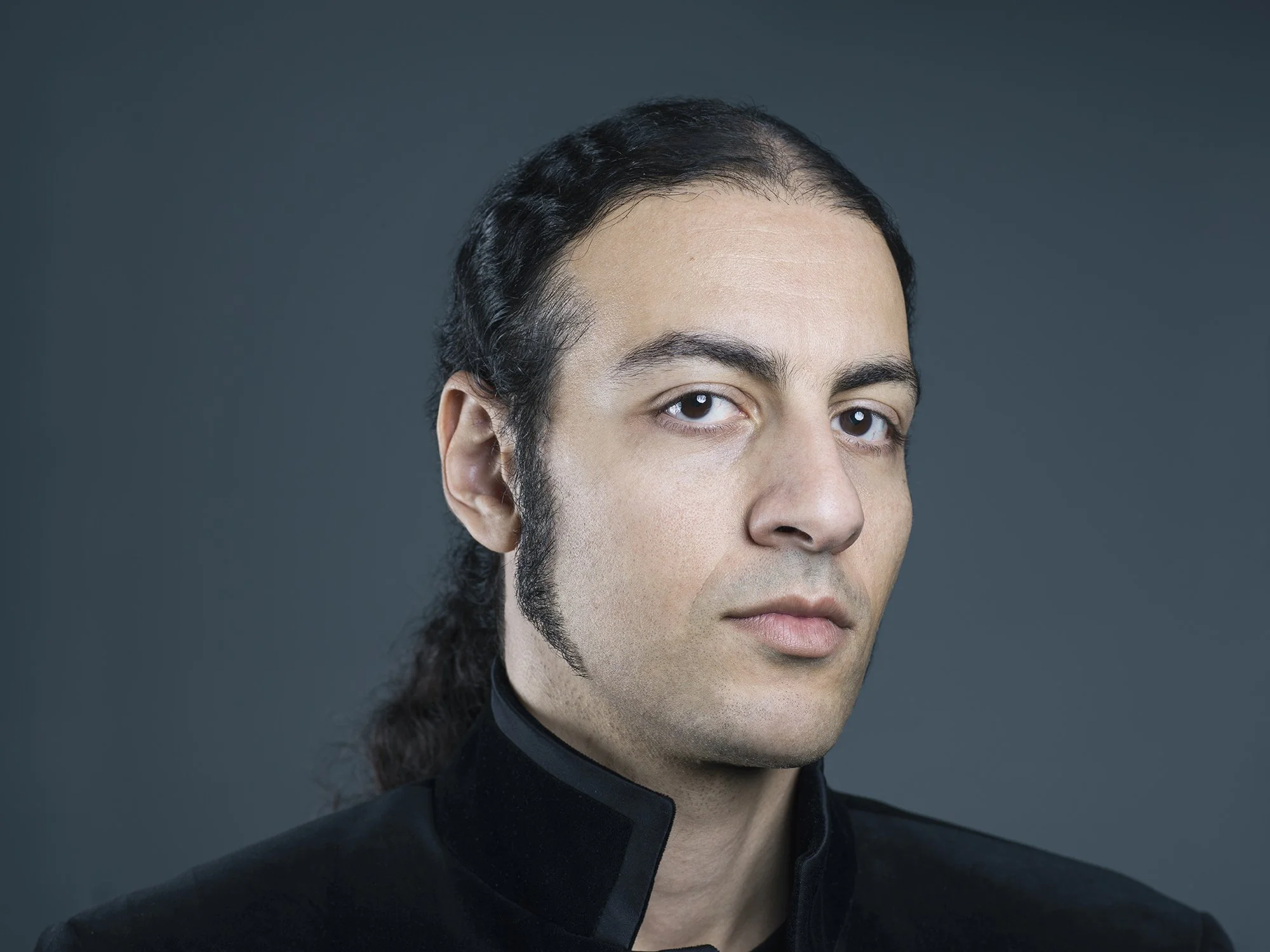 Portrait of a man with long dark hair, light skin, wearing a black high-collared shirt, against a plain dark background.