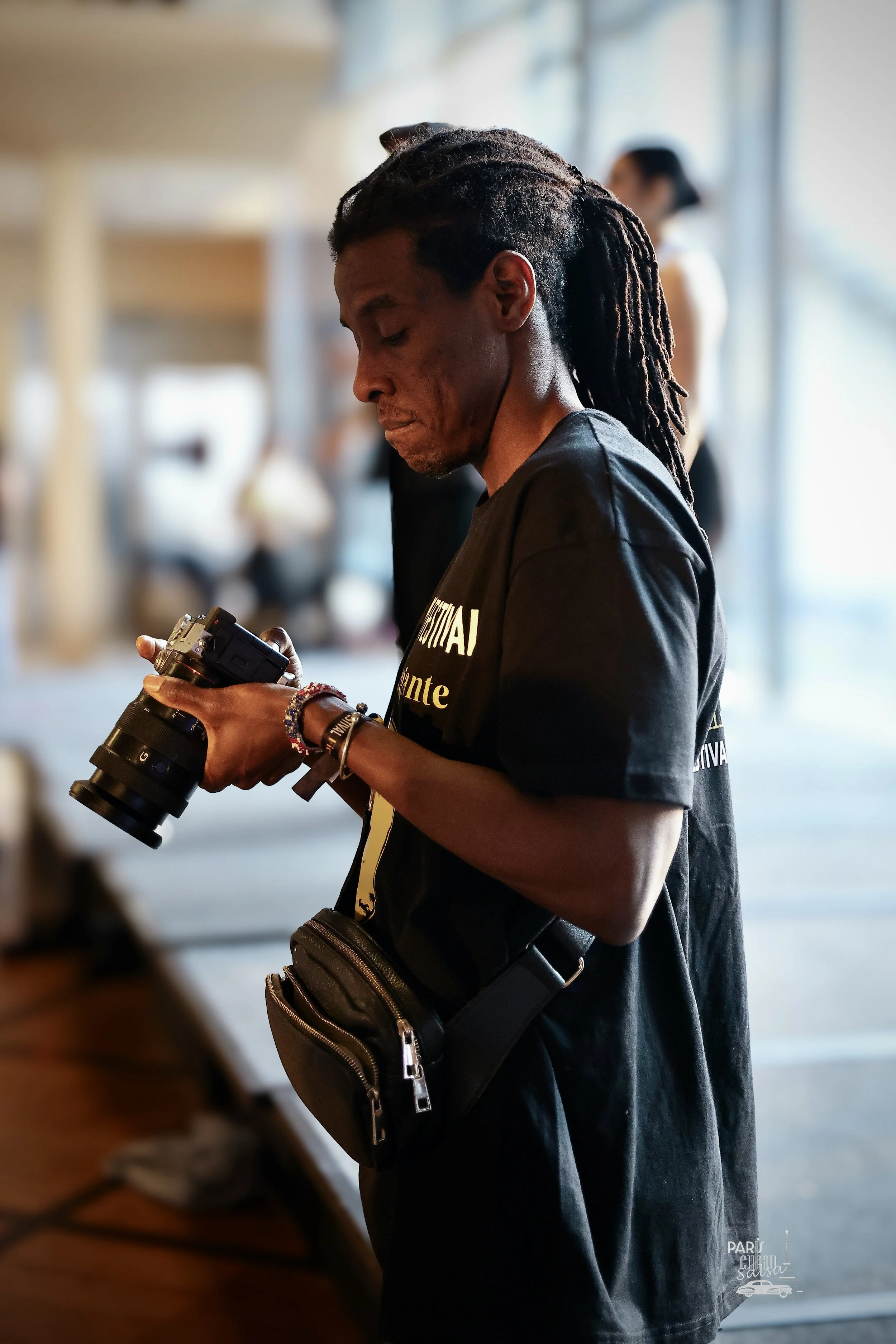 A man with dreadlocks wearing a black T-shirt and a small black bag is looking at a camera he is holding, standing indoors near a window with natural light.