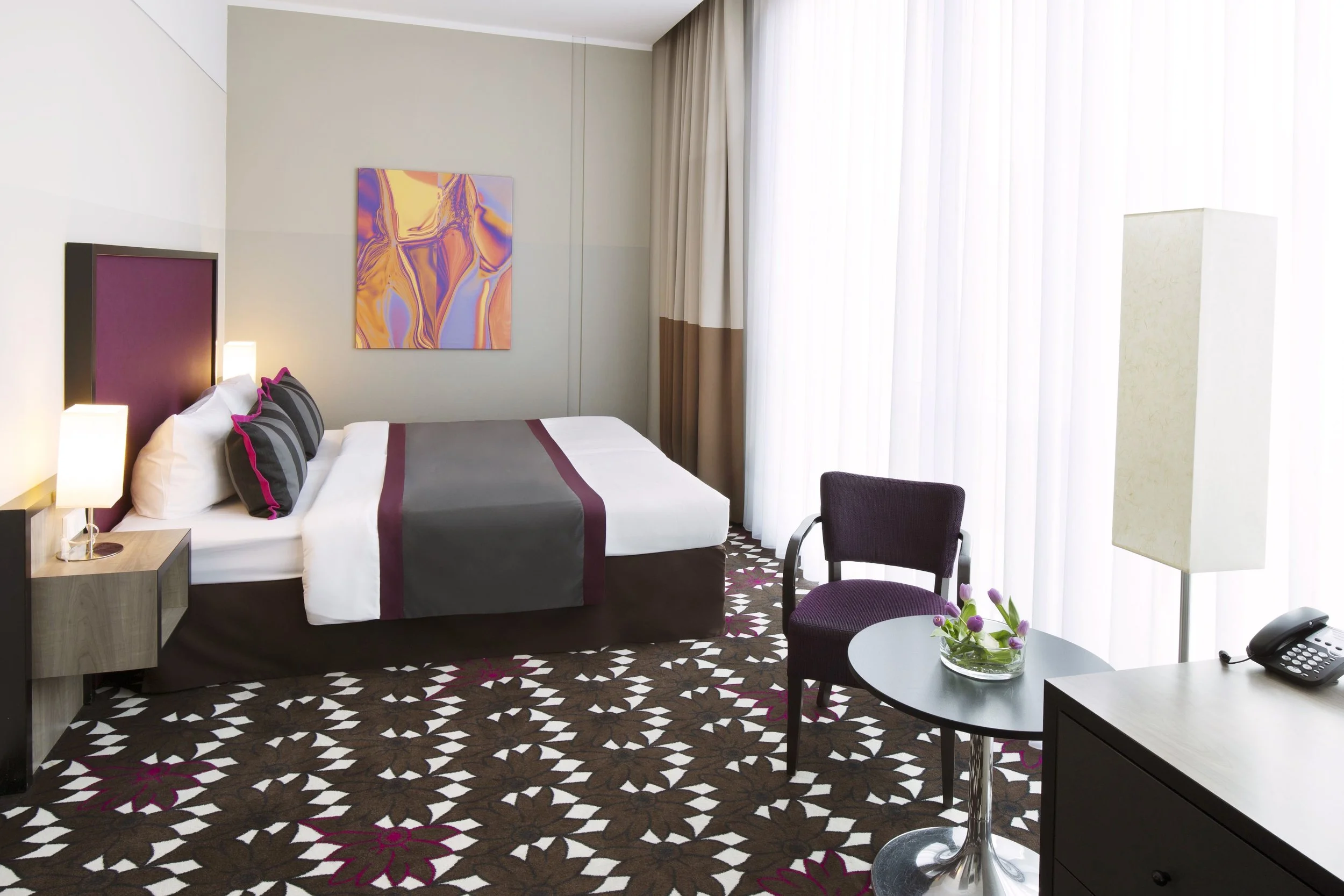 A hotel room with a neatly made bed featuring black, grey, and pink striped pillows, a nightstand with lamps, artwork on the wall, a patterned carpet, a small round table with a purple chair, a plant in a glass bowl, and large white curtains covering a window.