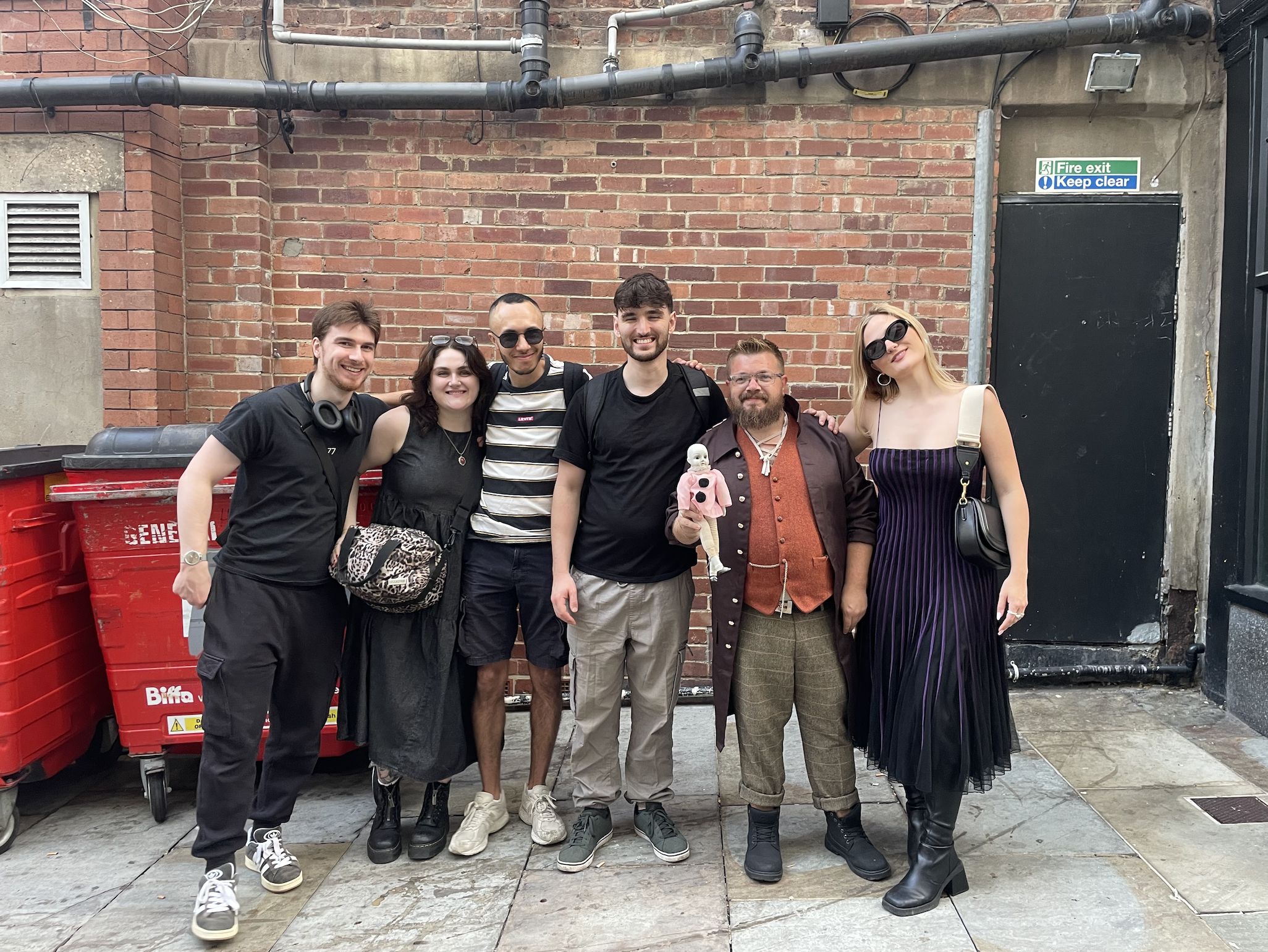 Group of six people standing side by side in front of a brick wall, smiling at the camera. They are dressed casually, with some wearing sunglasses and accessories, and one person is holding a small doll.