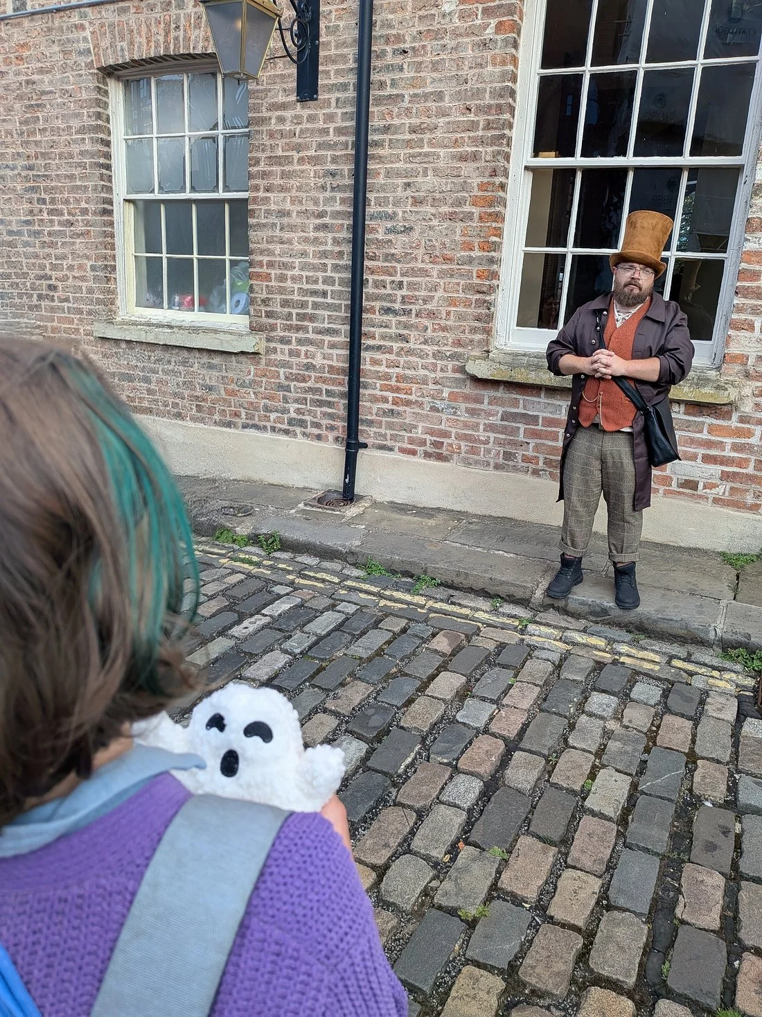 A person with long hair, wearing a purple sweater and a backpack, holding a small ghost-shaped plush toy, facing a man dressed in vintage-style clothing with a tall brown hat, standing on a cobblestone street outside a brick building.