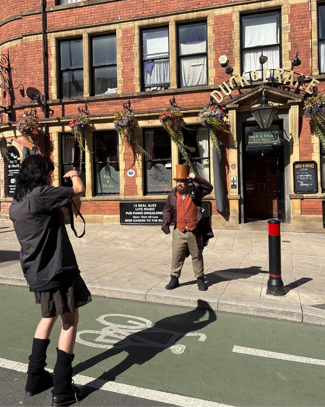 A man in vintage clothing with a top hat and beard stands on the sidewalk in front of a brick pub called Duck & Drake, posing for a photo being taken by a woman with a camera. The pub has hanging flower baskets and a sign advertising live music.