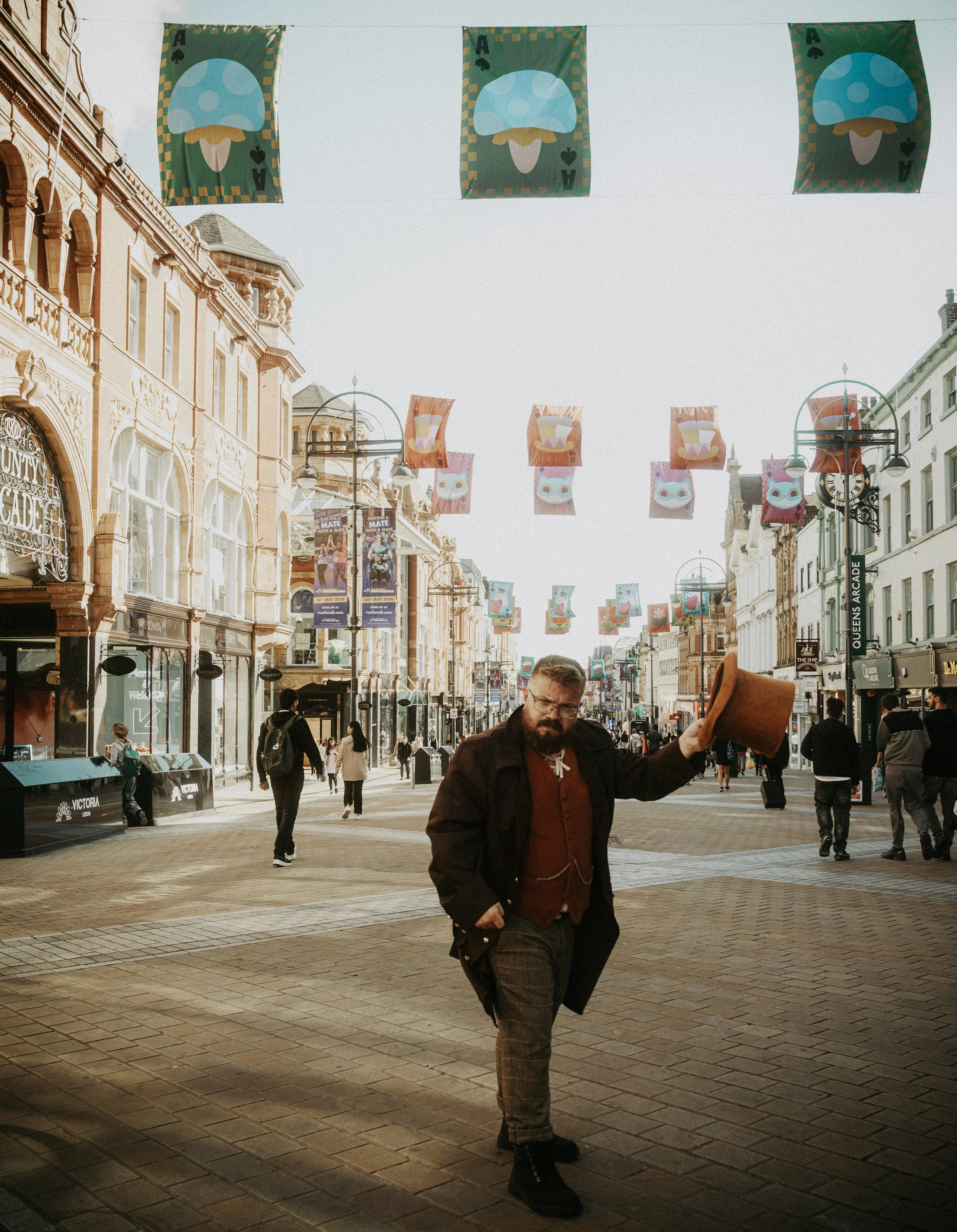A man with a beard and glasses standing on a busy city street, holding a hat, with colorful banners and historic buildings overhead.