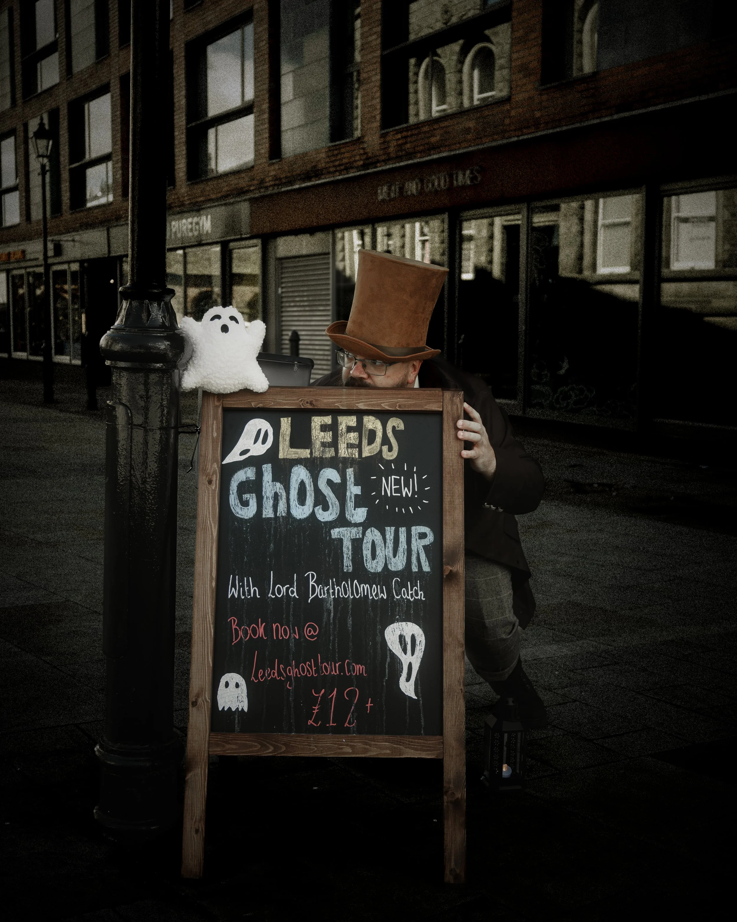 A man wearing a tall brown top hat crouches behind a chalkboard sign on a city sidewalk. The sign promotes a Leeds Ghost Tour with Lord Bartholomew Catch, displaying cartoon ghost illustrations and reservation information. A small plush ghost is atta