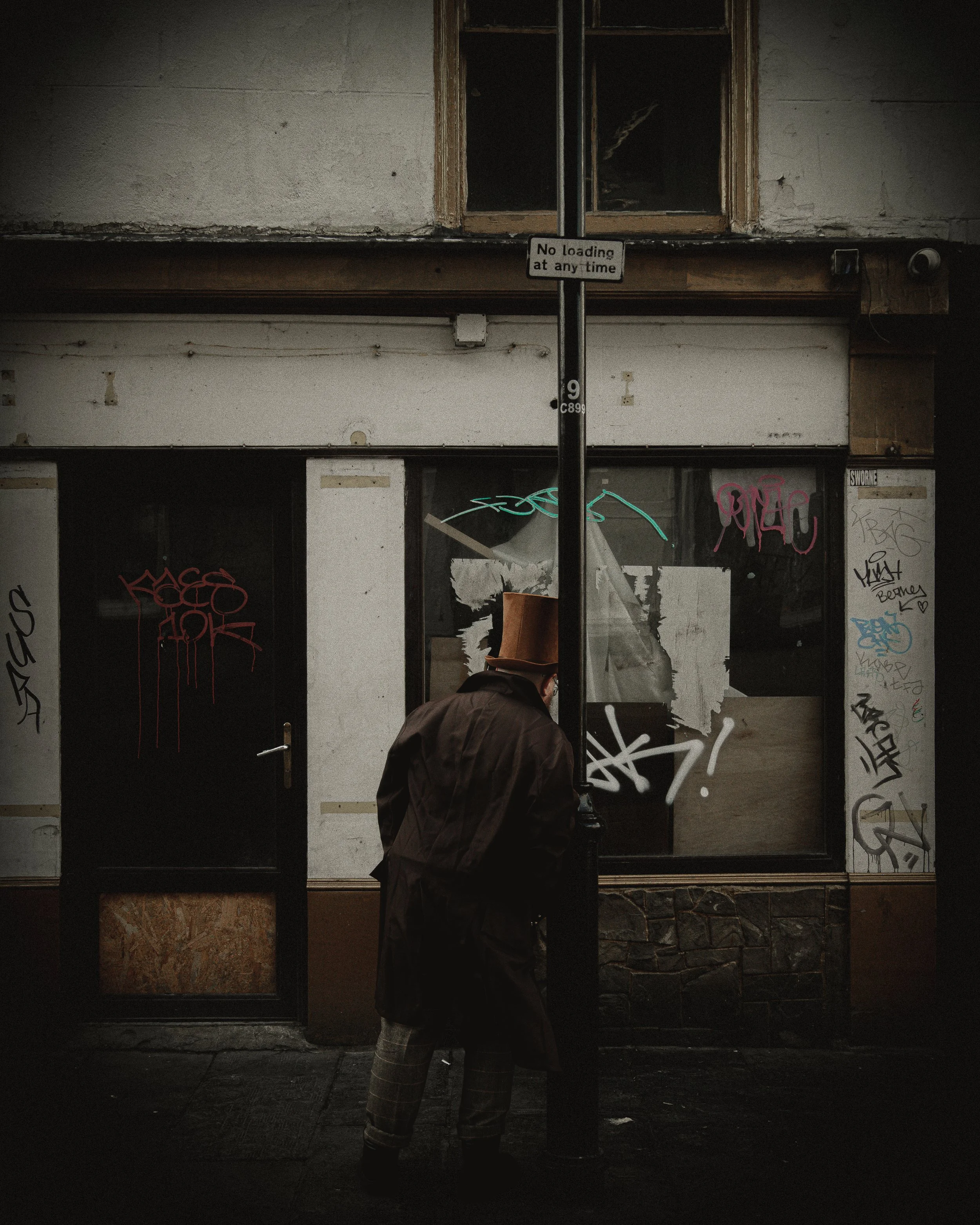A person wearing a brown top hat and dark coat standing on a dark city sidewalk, leaning against a black pole with a sign that says 'No loading at any time.' Behind him is a building with boarded-up windows and graffiti on the walls.