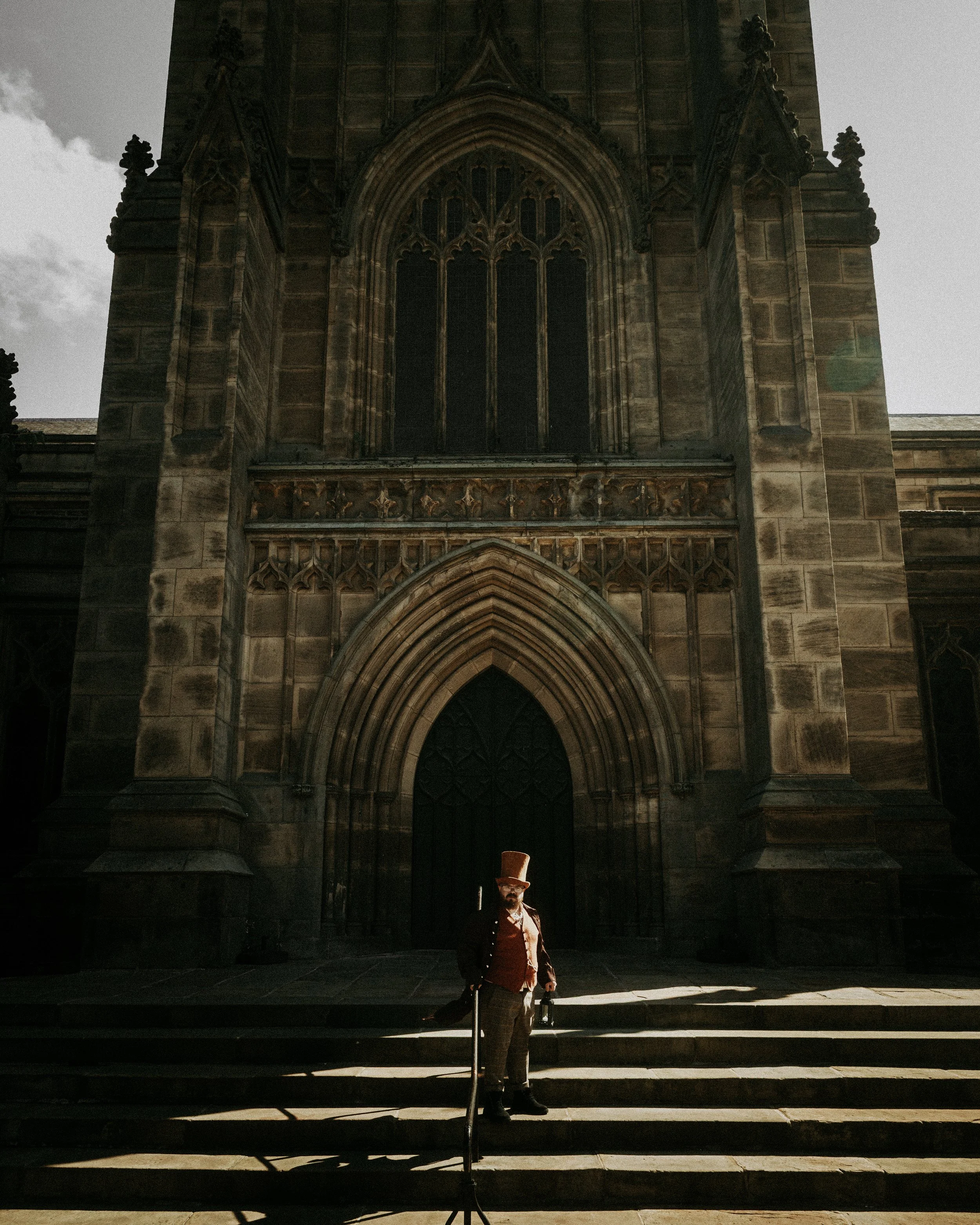 A man dressed in historical attire, including a tall top hat, standing on the steps in front of a large, Gothic-style cathedral.