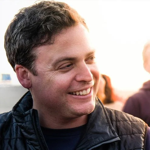 Smiling man wearing a black Patagonia vest outdoors.