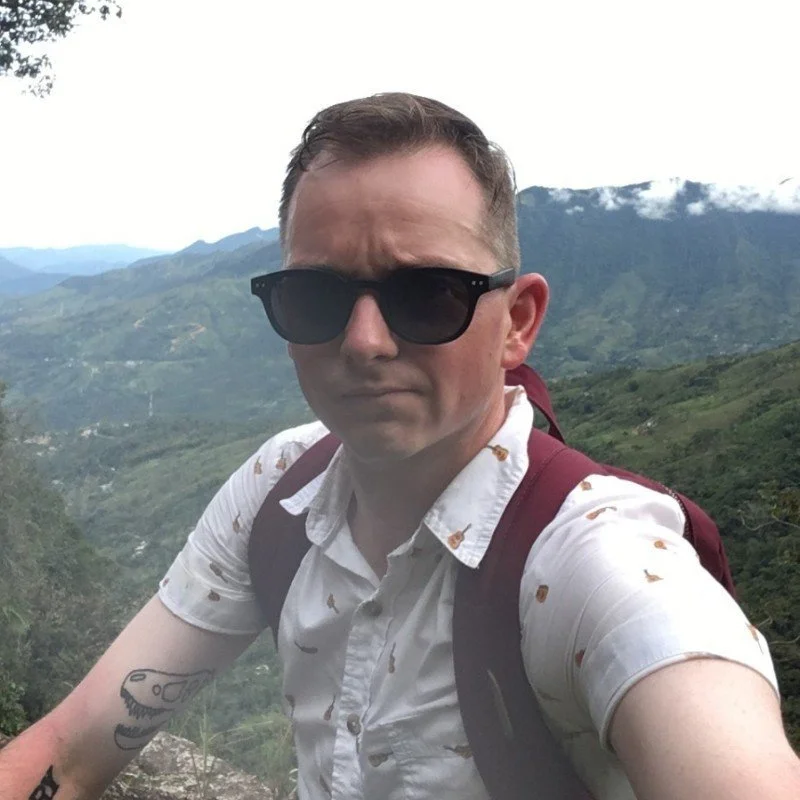 Person wearing sunglasses and a patterned shirt, standing in front of a mountainous landscape.