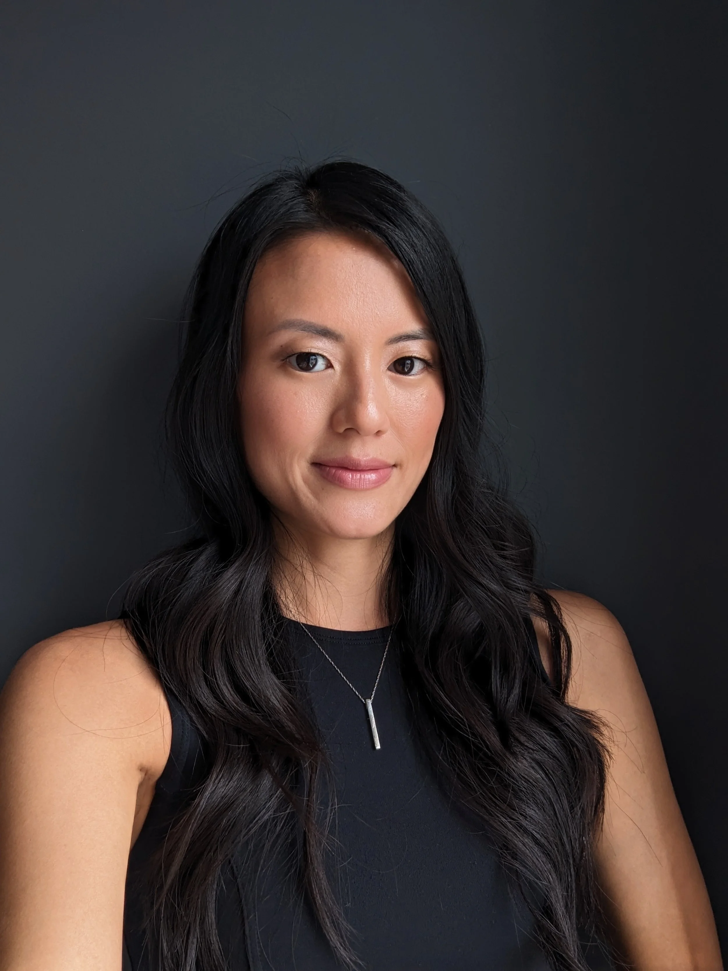 Person with long black hair wearing a black sleeveless top and a necklace, standing against a dark background.