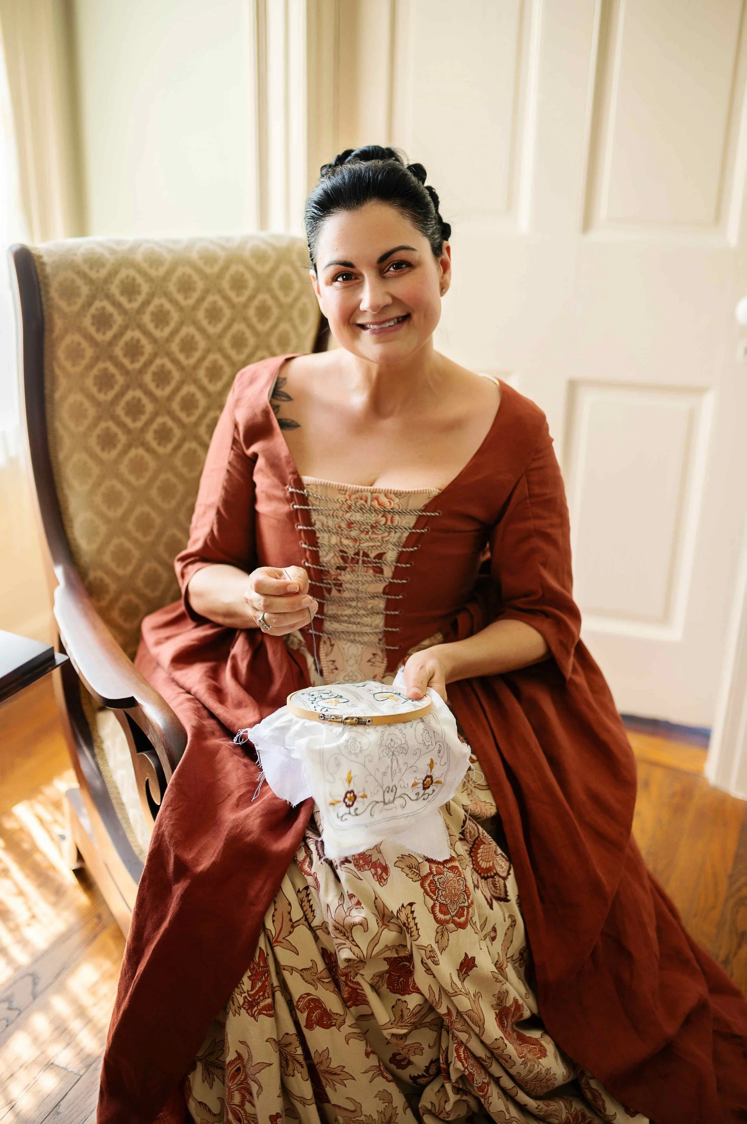 A woman is sitting on a chair, smiling, and working on hand embroidery with a hoop. She is wearing a Colonial-style dress with floral patterns and a brown outer garment. The room has warm lighting with cream-colored walls and wooden flooring.