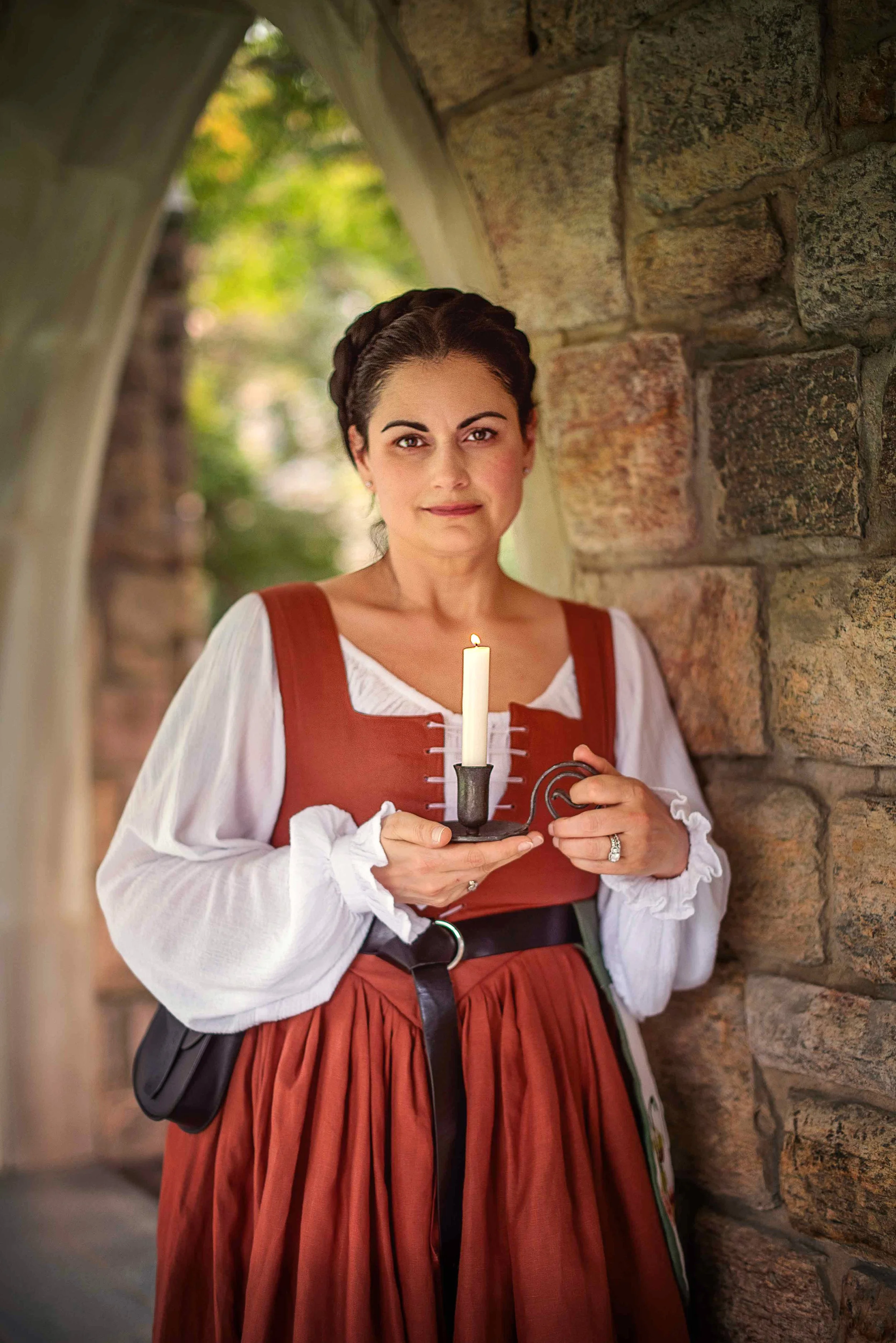 A woman dressed in a traditional medieval kirtle holding a candle in a holder, standing against a stone wall under a stone arch, with colorful leaves visible in the background.