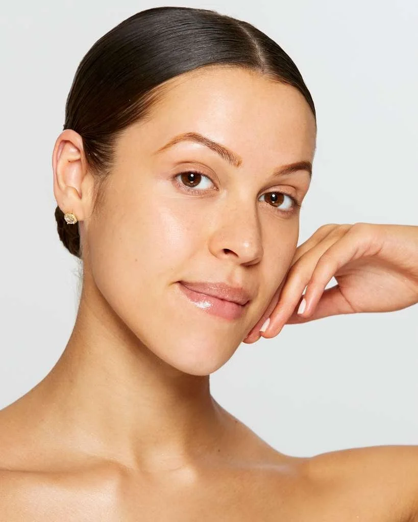 A close-up headshot against a light gray background. Her hair is pulled back in a sleek low bun, and she rests her chin lightly on the back of her hand. The framing is tight, from the shoulders up, with soft natural lighting.