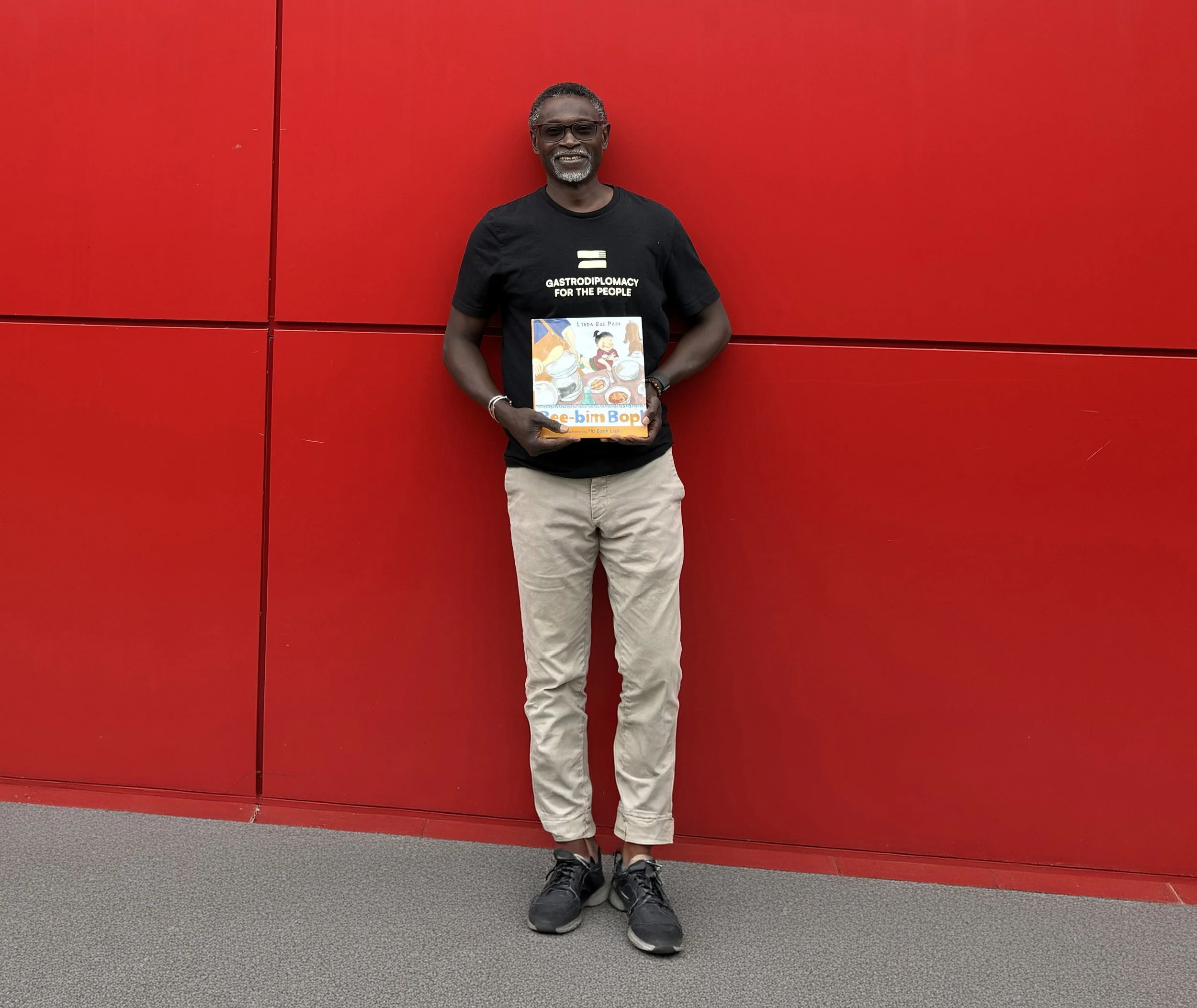 Chef Pierre poses in front of a bright red wall holding a children's book from a volunteer session