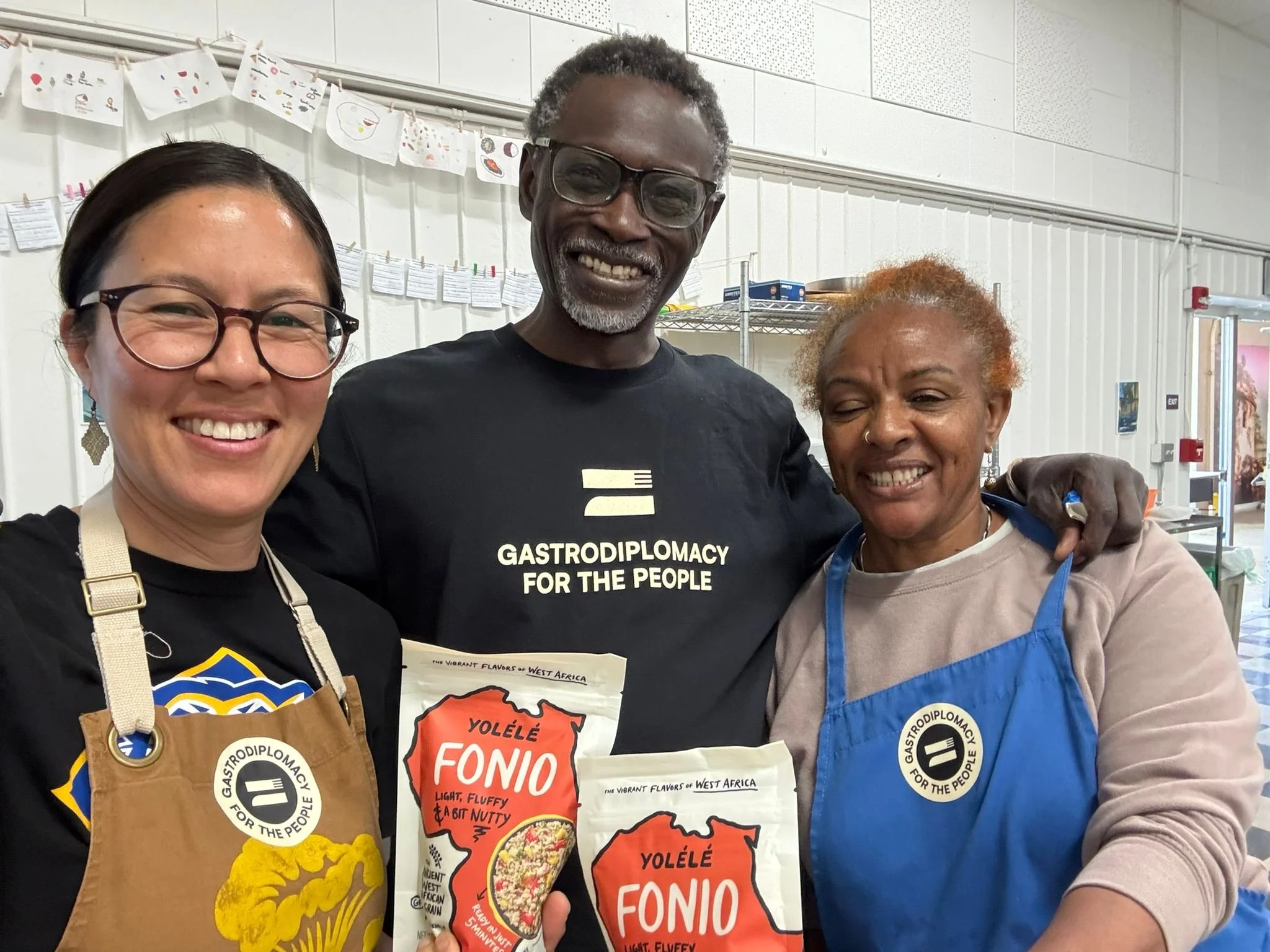 Chef Pierre poses with two Oakland USD staff in aprons holding bags of Fonio grains and smiling