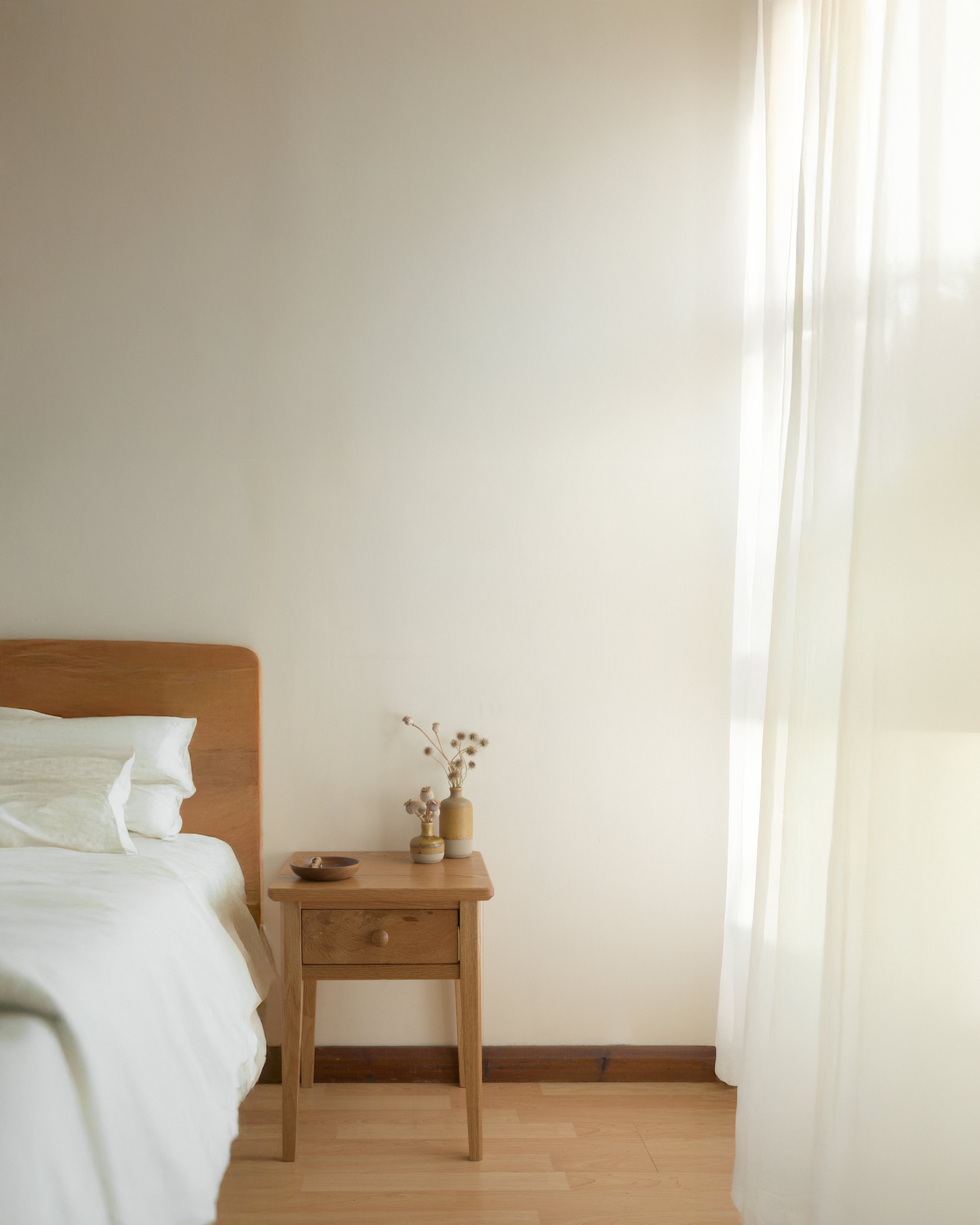Part of a bedroom with a wooden bed, white bedding, a small wooden nightstand with vases and dried flowers, and cream curtains next to a window.