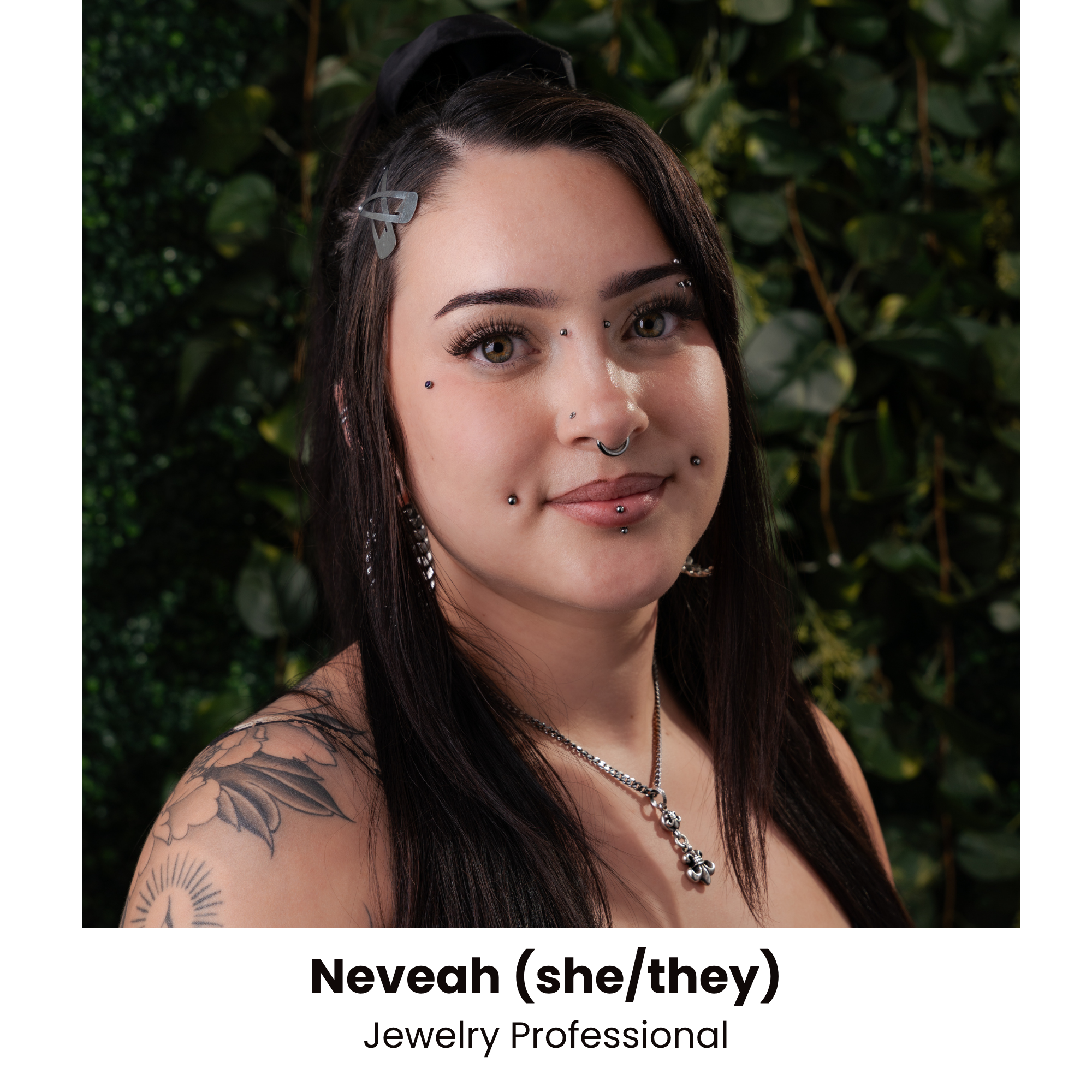 Close-up of a young woman with multiple facial piercings, tattoos on her arm, long dark hair, and wearing a necklace and hair clips, standing outdoors with blurred green foliage in the background.