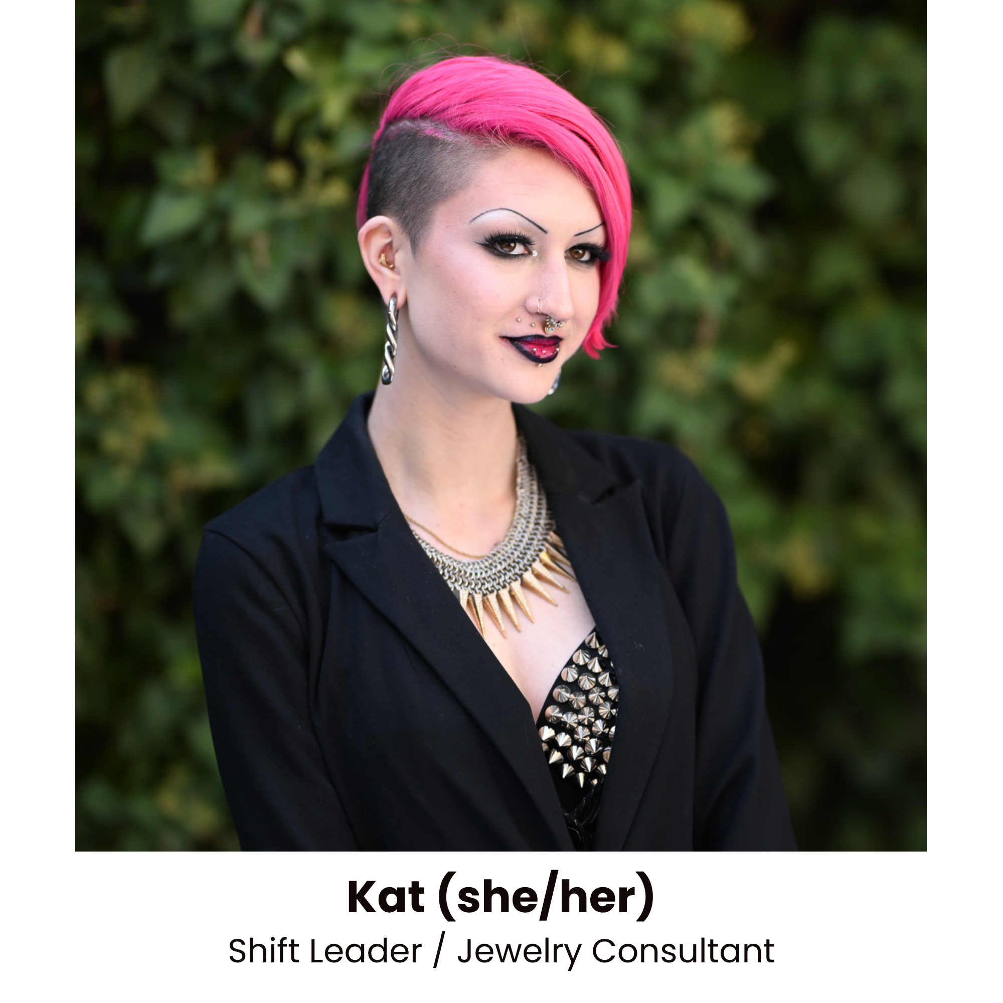 A woman with pink hair styled in an edgy undercut, wearing makeup with dark eyeliner and lipstick, accessorized with earrings and a statement necklace, standing outdoors with greenery in the background.