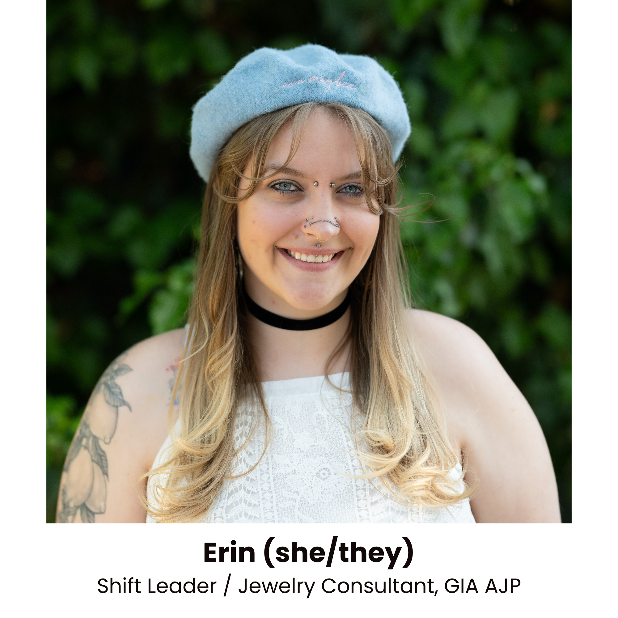 Close-up of a smiling young woman with long wavy blonde hair, multiple facial piercings, blue eyes, wearing a light blue beret with pink embroidered writing, a black choker, and a white sleeveless dress, with a green foliage background.