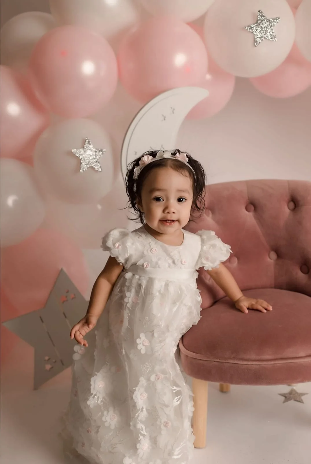 Toddler in a white dress standing by a pink chair, surrounded by pink and white balloons with star decorations.
Everly Everafter Kids | Children's Clothing Store