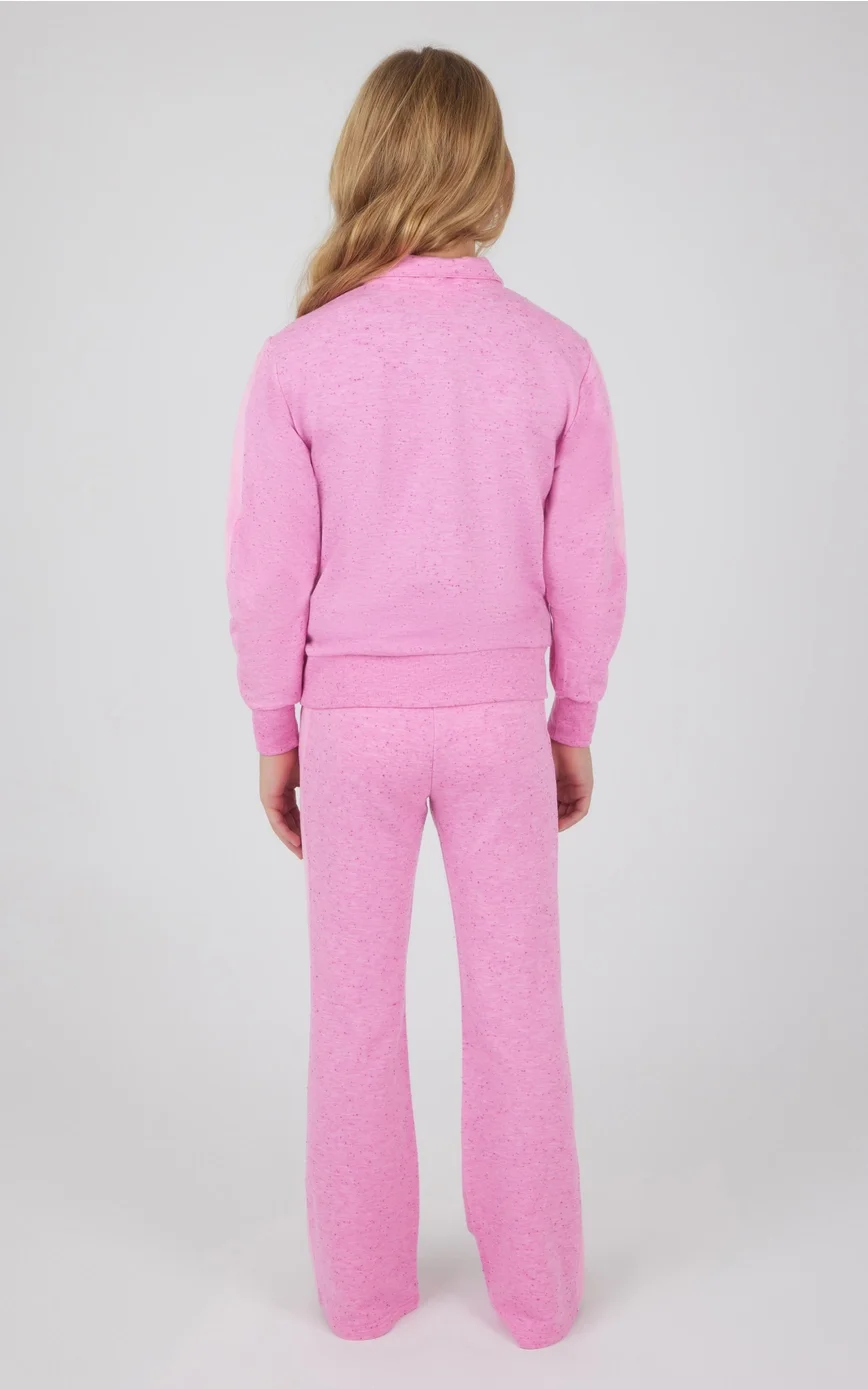 Back view of a woman with long blonde hair wearing a pink speckled loungewear set consisting of a sweatshirt and matching sweatpants, standing against a plain white background.